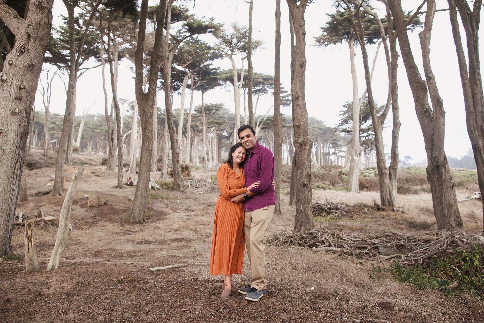 A couple in vibrant orange and maroon clothing stands arm-in-arm among towering cypress trees, smiling warmly at the camera with dappled light filtering through the canopy