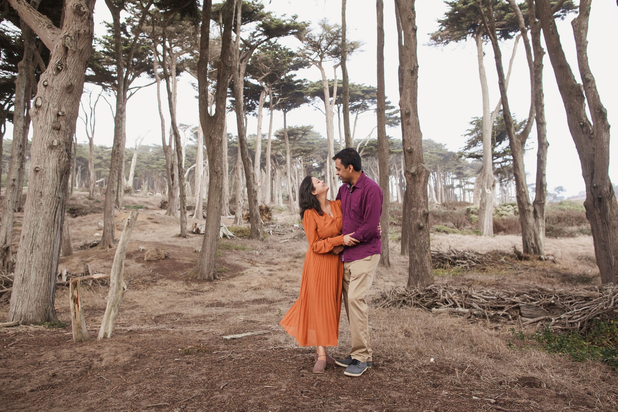 A couple in vibrant orange and purple clothing stands beneath towering cypress trees, gazing at each other with warmth amid dappled forest light