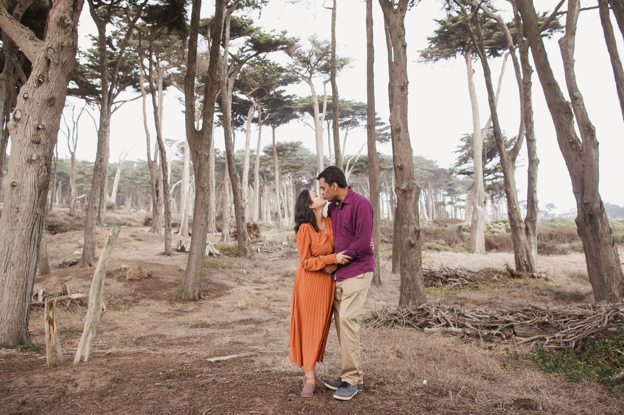 A couple in vibrant orange and burgundy clothing shares an intimate moment among towering cypress trees on sandy forest ground