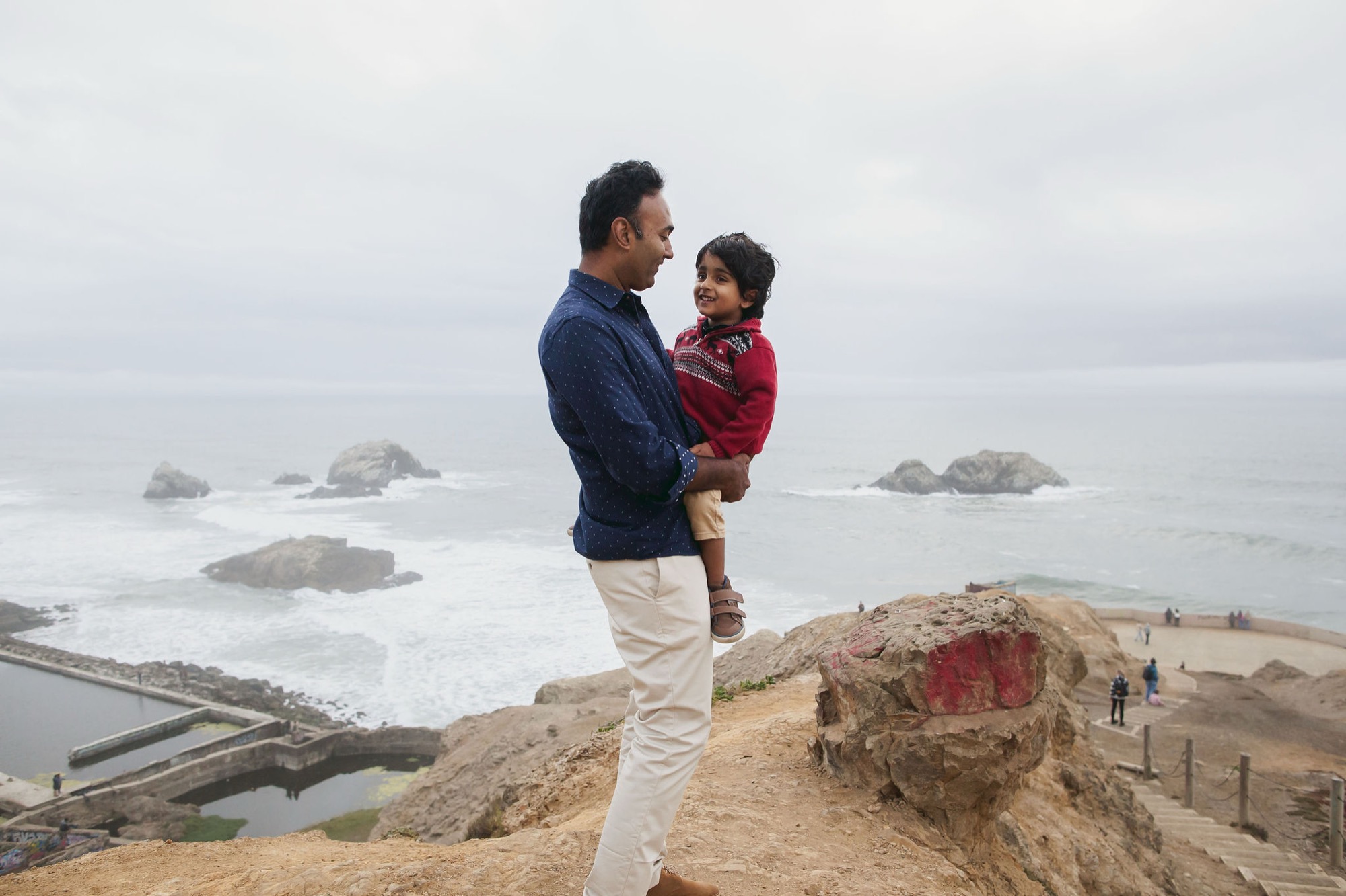 A father holds his young son while gazing toward the ocean, standing on a windswept coastal bluff with offshore rocks visible in the misty background