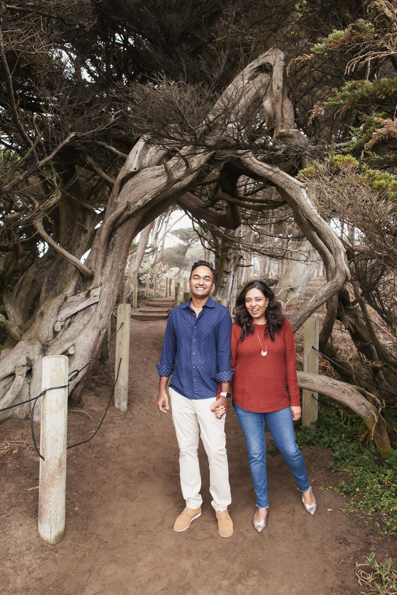 A couple holds hands while smiling beneath a gnarled, twisted cypress tree on a dirt trail, with fog-shrouded cypress avenue stretching behind them