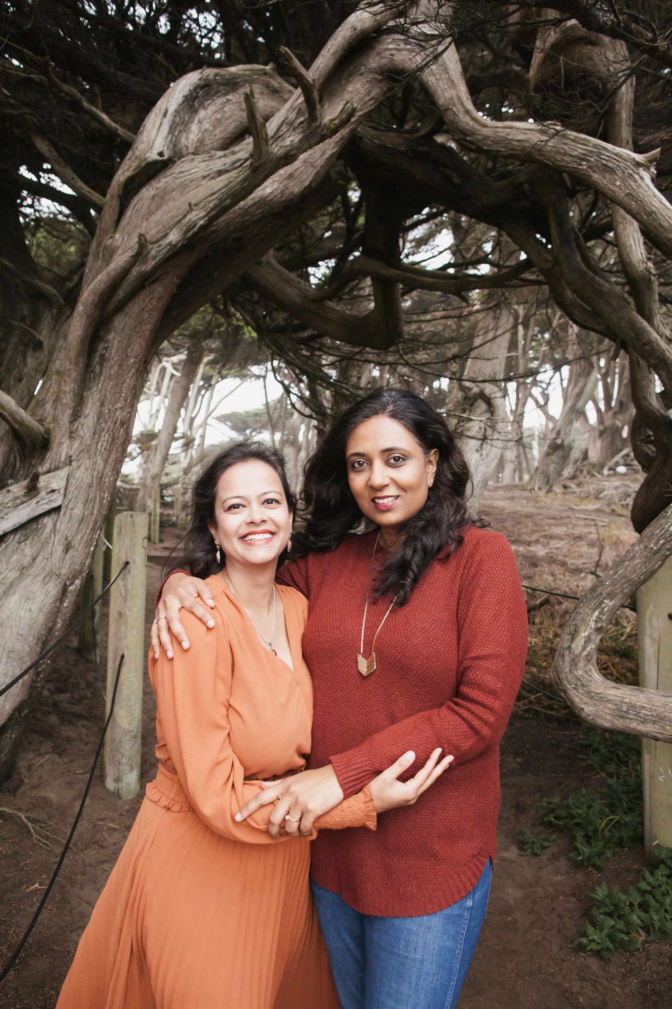 Two women in warm earth tones embrace beneath gnarled cypress branches, their genuine smiles framed by twisted wood and coastal light