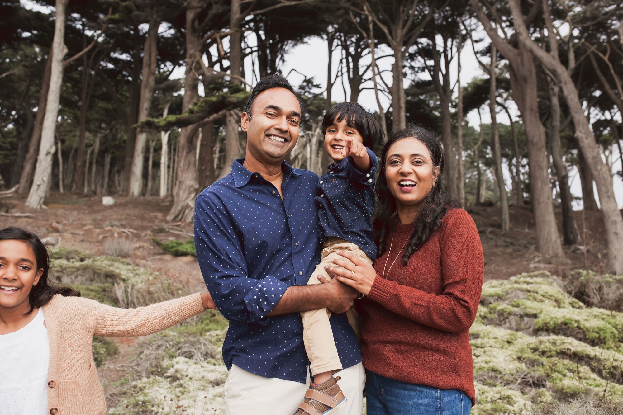 A joyful family of four laughs together among towering cypress trees, with the father holding their young son while the mother beams beside them