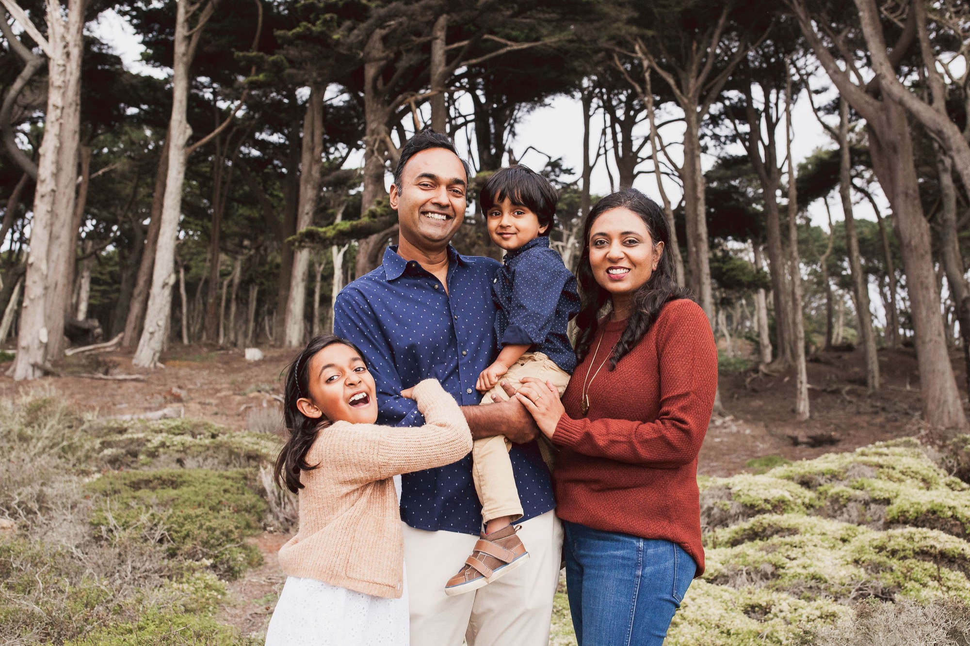 A joyful family of four laughs together among towering eucalyptus trees, their warm clothing and genuine smiles radiating happiness in dappled woodland light