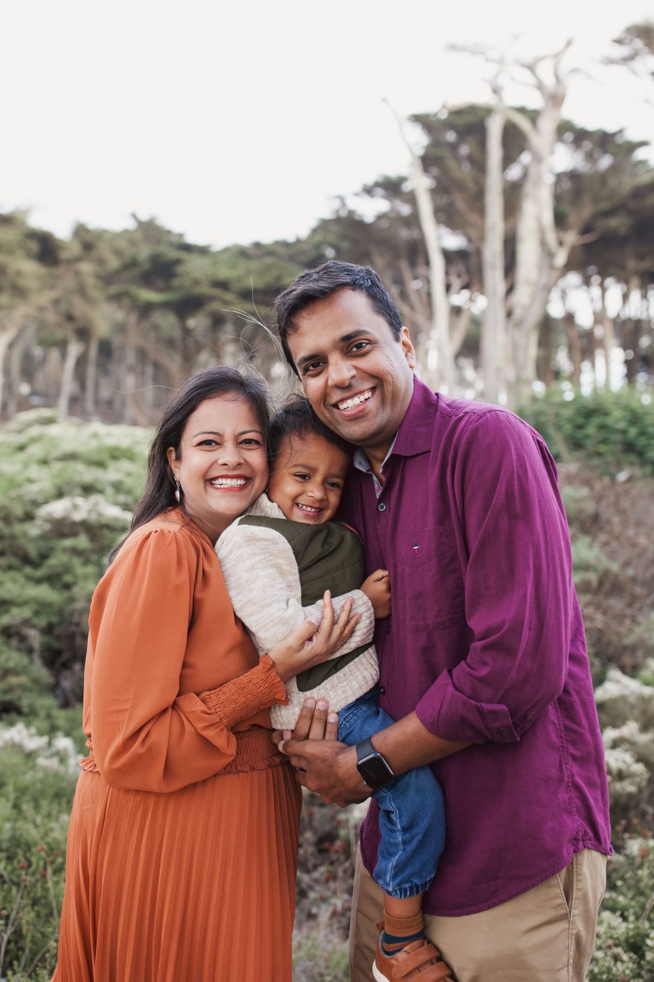 A smiling family of three poses together against windswept cypress trees and coastal vegetation at Land's End in San Francisco