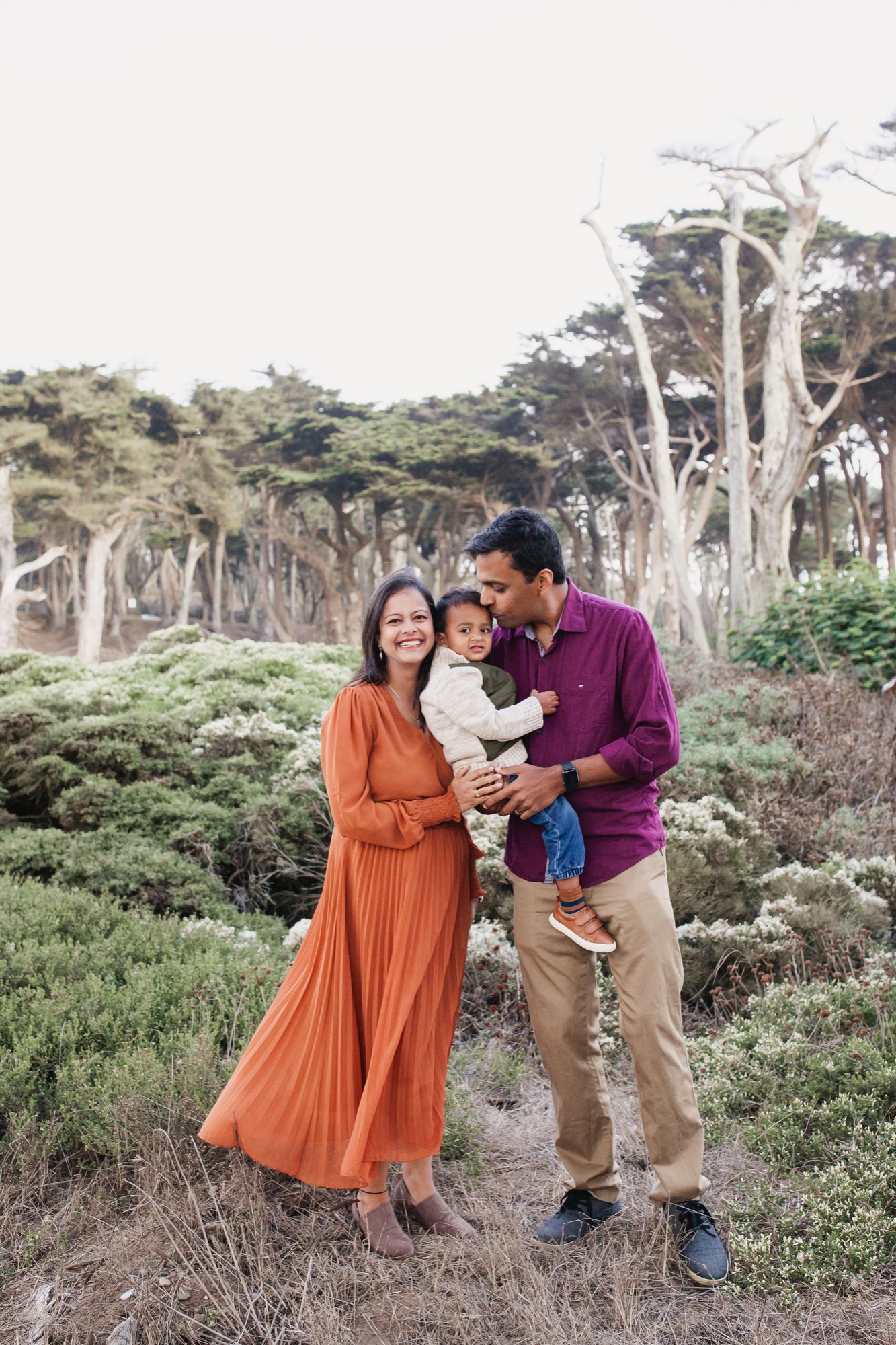 A family of three stands together on a dusty trail surrounded by coastal scrub, with distinctive wind-swept cypress trees towering behind them in soft afternoon light