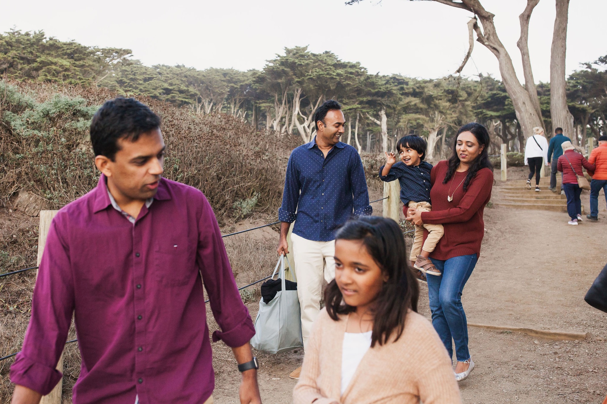 A multi-generational group walks a tree-lined coastal path, with a man in maroon leading while others follow in casual conversation under overcast light