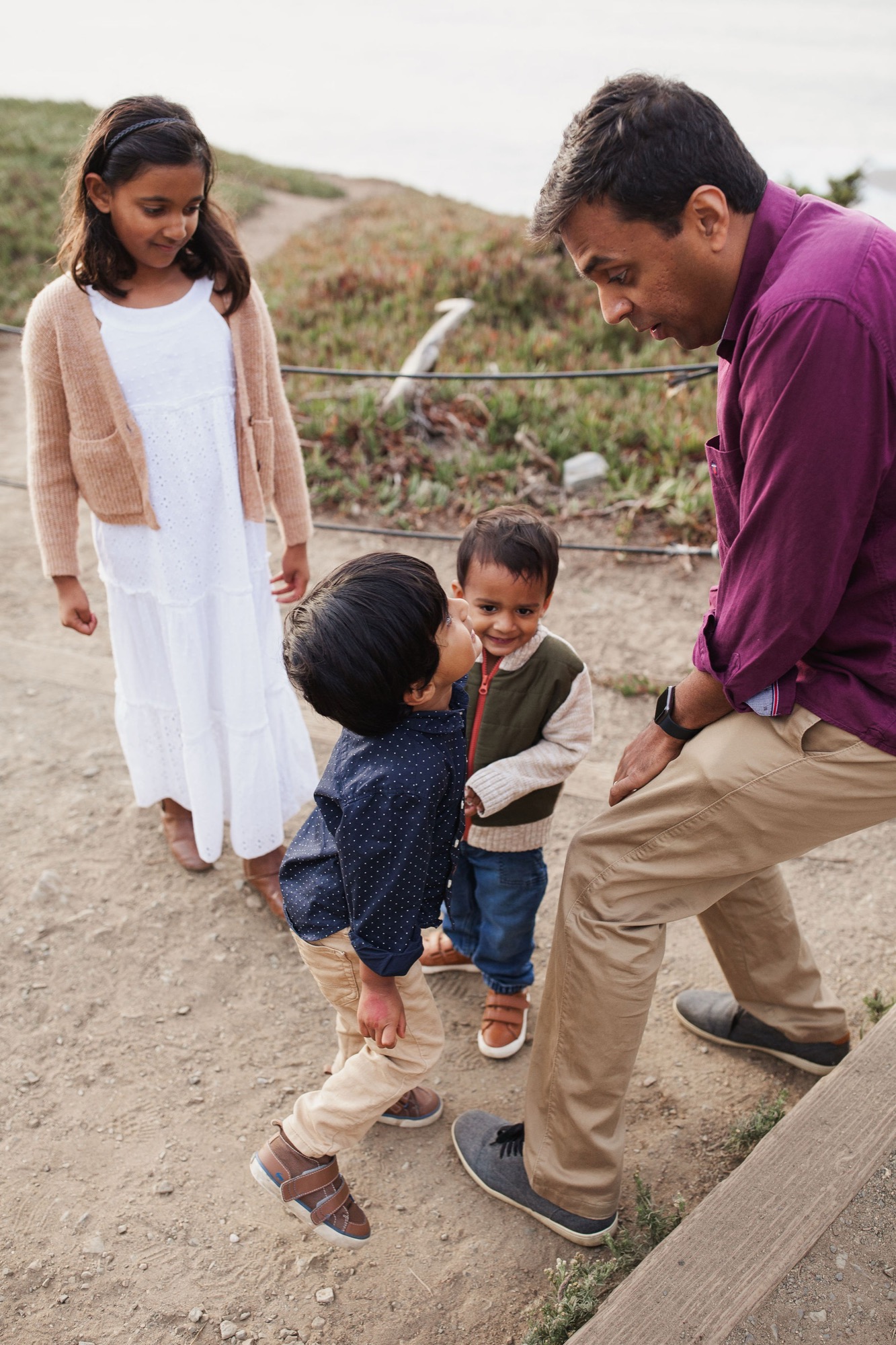 A father in purple kneels to engage with his young daughter while older siblings watch nearby on a dusty agricultural path