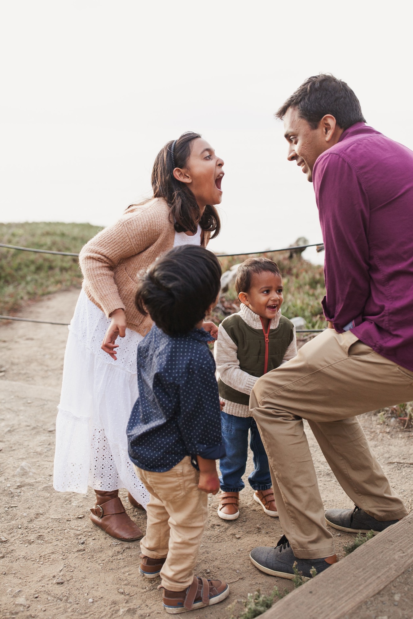 Parents and two young children laugh together on a dusty trail, their genuine joy captured against an overcast coastal sky and green hillside