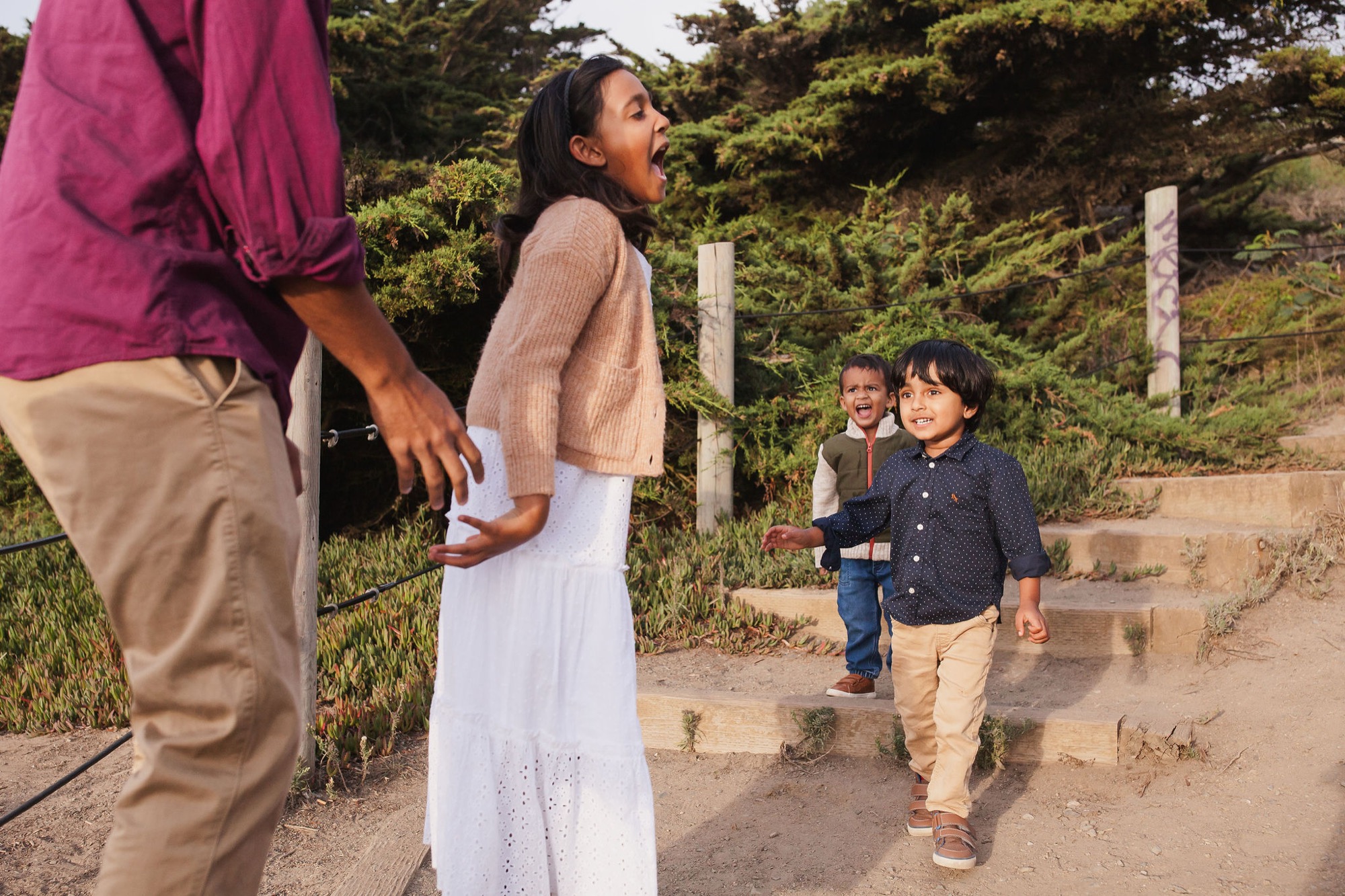 Two young boys in bright shirts run toward their parents on a dusty coastal trail, their genuine joy captured mid-stride