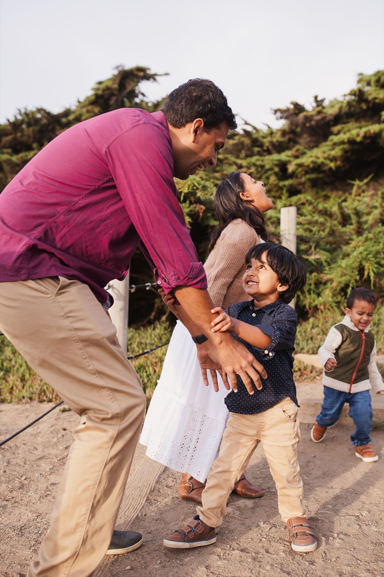 Father bends down to engage with his young son on a dusty trail, while mother and siblings look on with genuine smiles amid windswept coastal vegetation