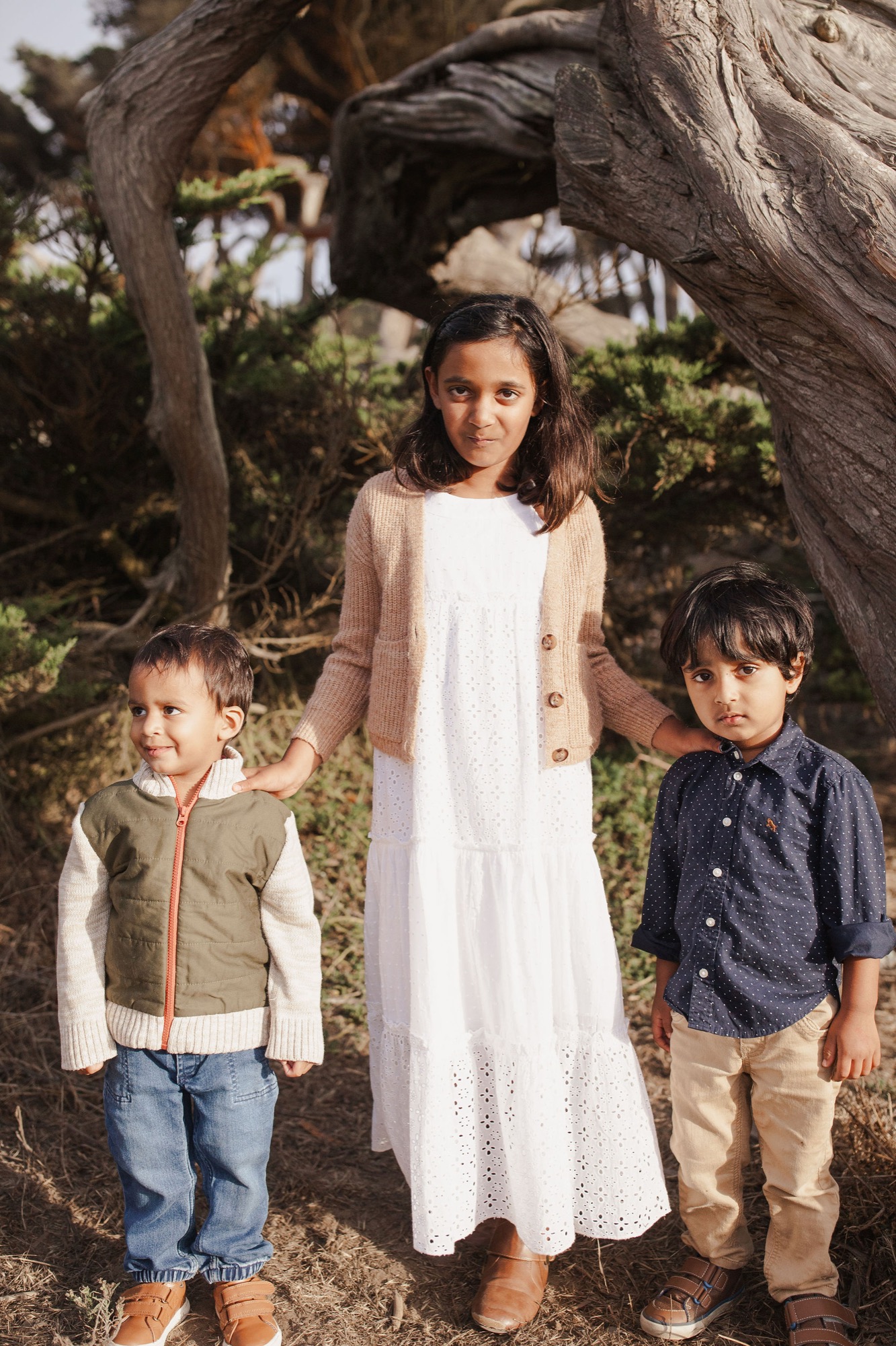 Three children stand framed between gnarled tree trunks, the older girl in white eyelet gazing directly at the camera while her younger brothers lean into her sides