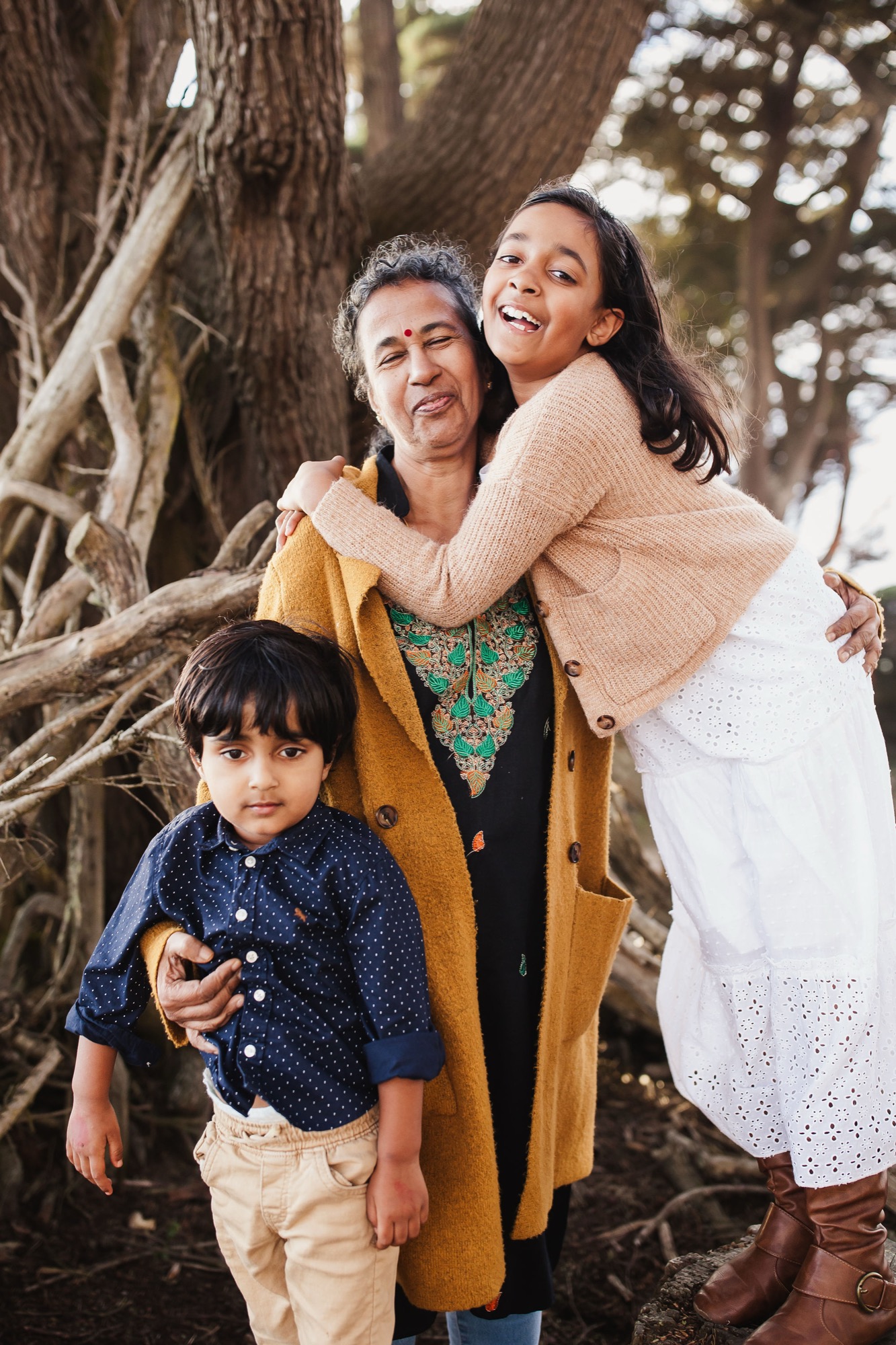 An intergenerational family shares genuine laughter beneath towering coastal redwoods, their warm-toned layers complementing dappled afternoon light filtering through ancient trees