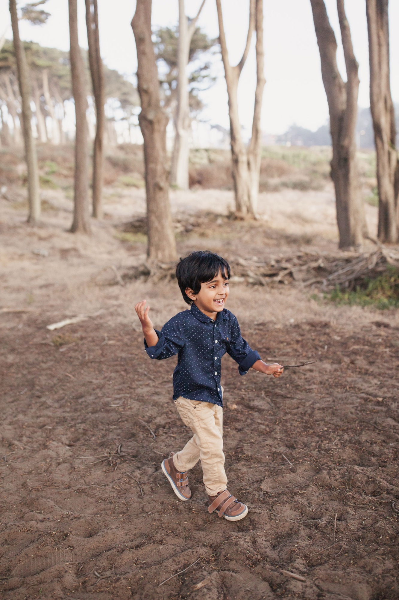A young boy with dark hair runs joyfully through a eucalyptus grove, arms open wide, smiling broadly as soft afternoon light filters through the tall trees