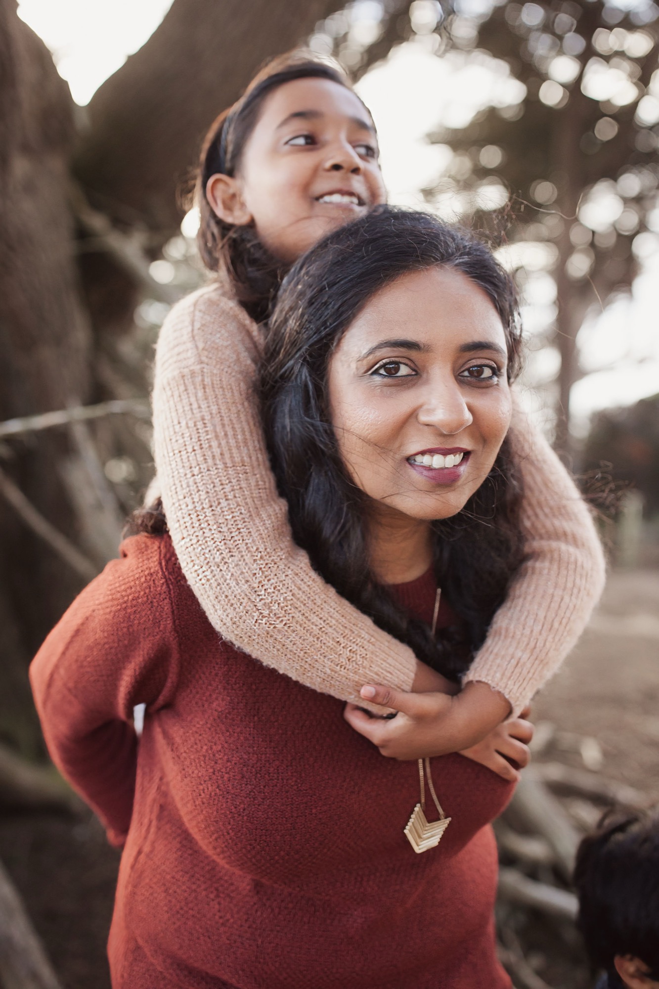 A woman in a rust sweater smiles warmly while her daughter perches on her shoulders beneath towering trees, golden afternoon light filtering through the canopy