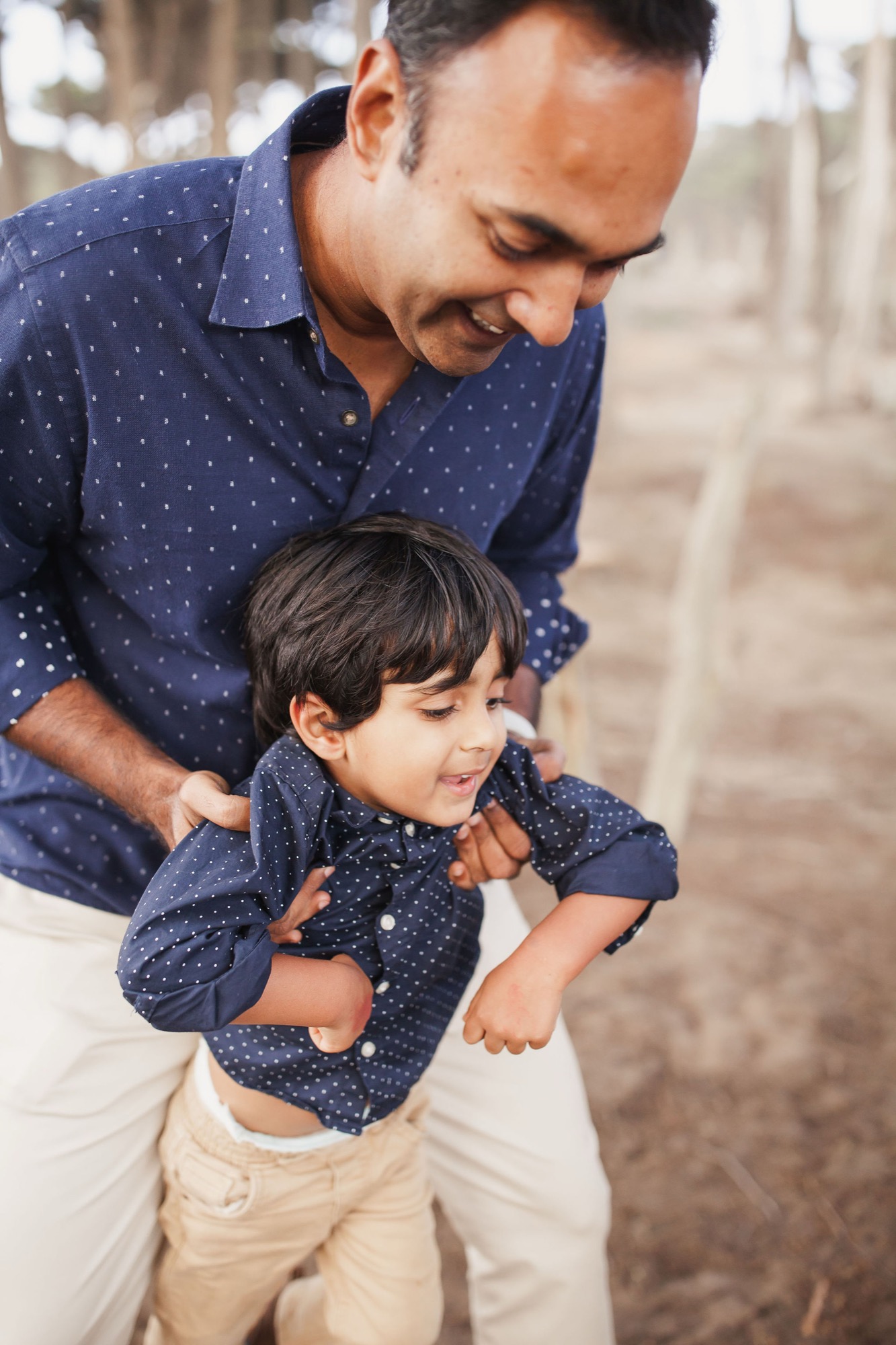 A father guides his young son along a coastal path, both wearing matching navy polka-dot shirts, sharing a genuine moment of connection and joy