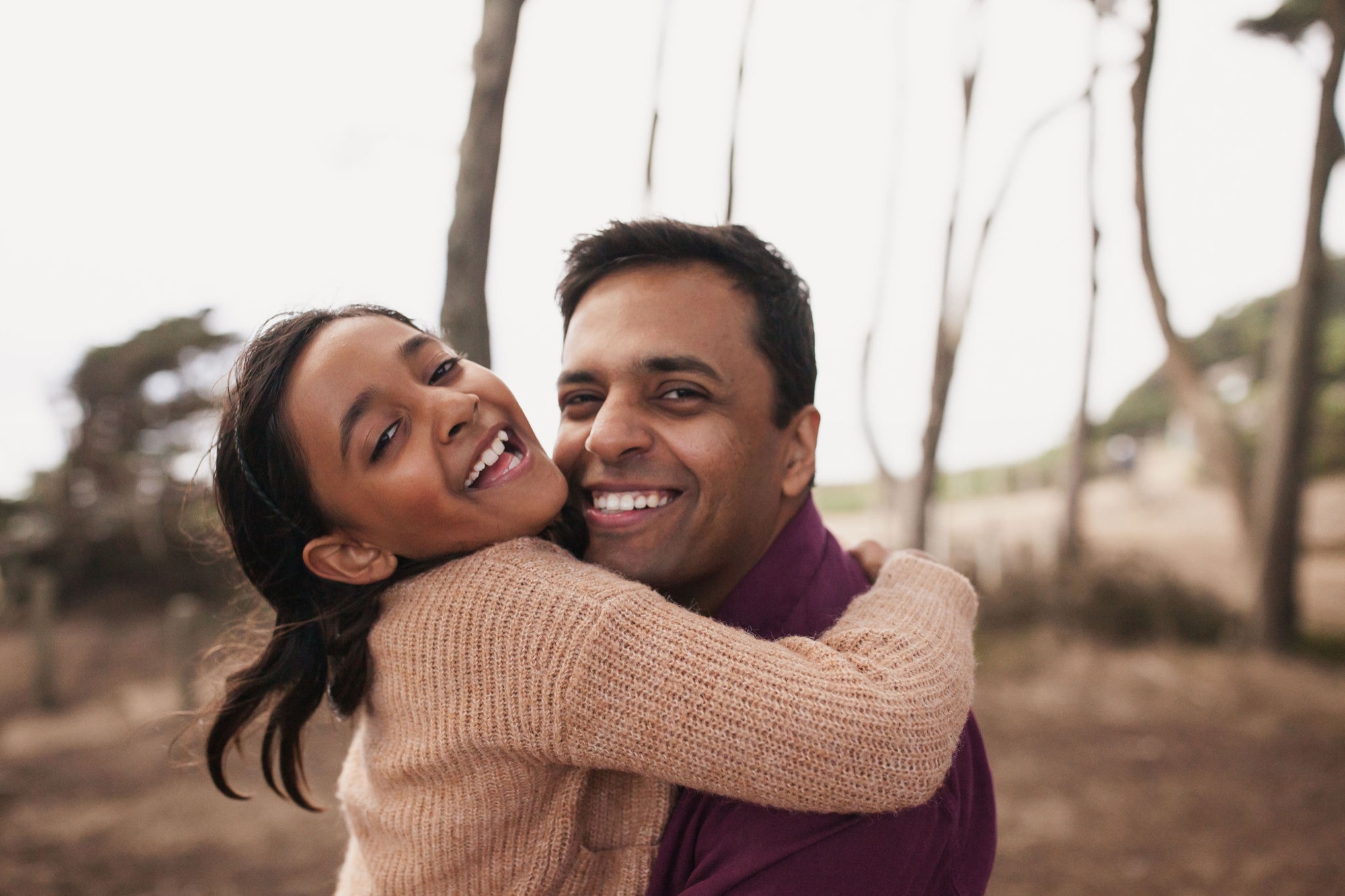 A couple laughs joyfully on a coastal trail, the man giving the woman a piggyback embrace with bare trees and misty hills behind them