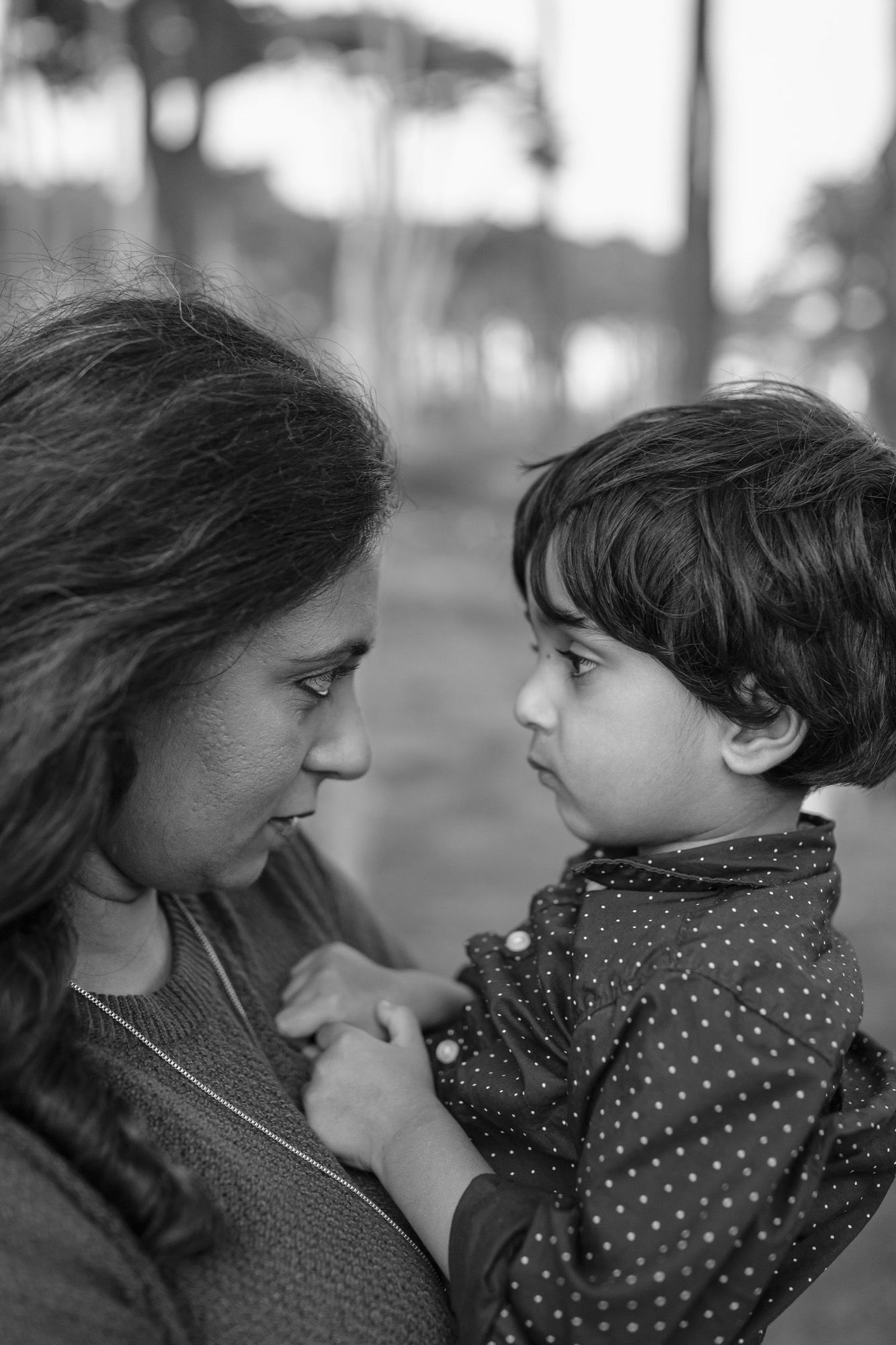 A mother and young child share an intimate moment in profile, their foreheads nearly touching as soft natural light catches their expressions against a blurred coastal backdrop