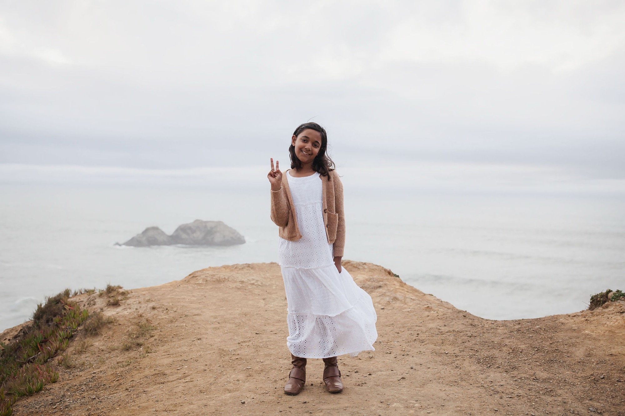 Young girl in white eyelet dress and tan cardigan makes a peace sign while standing on a sandy hilltop overlooking the ocean and distant rocky islands