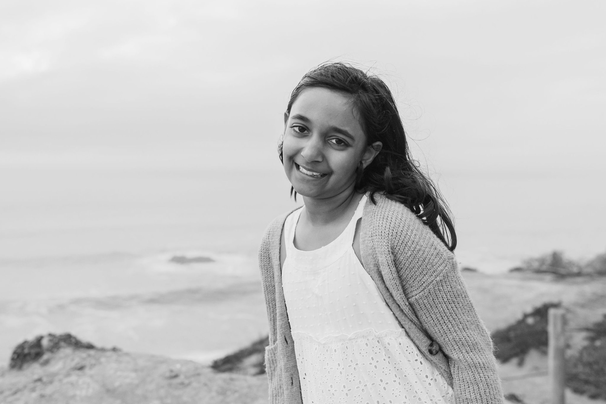 Young girl in cardigan smiles warmly at camera on misty coastal bluffs, ocean and distant hills softly blurred behind her