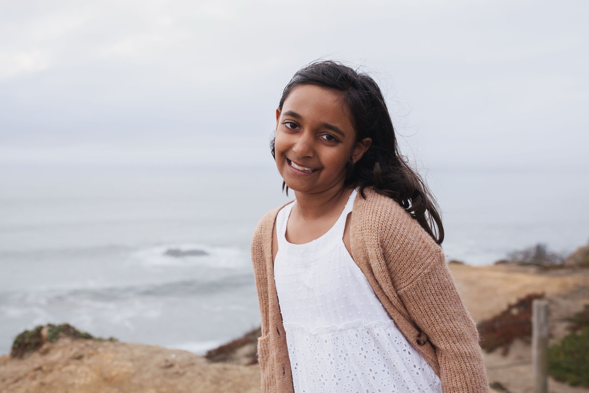 Young girl with long dark hair smiles warmly at camera on windswept beach, wearing cream cardigan over white top with ocean and overcast sky behind