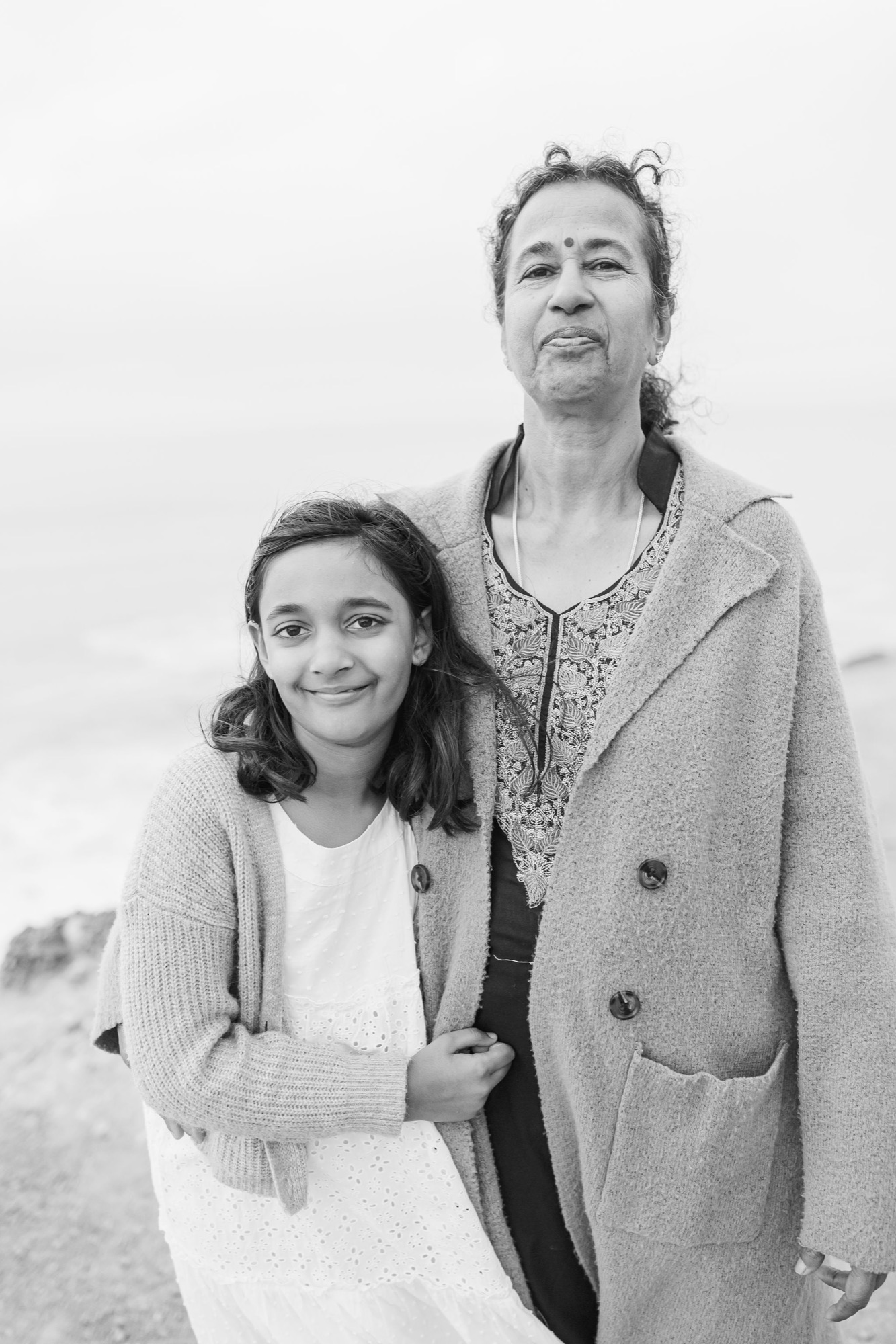 A woman and young girl stand close together on a misty coastal bluff, both wearing cardigan sweaters and gazing thoughtfully upward in soft, diffused light