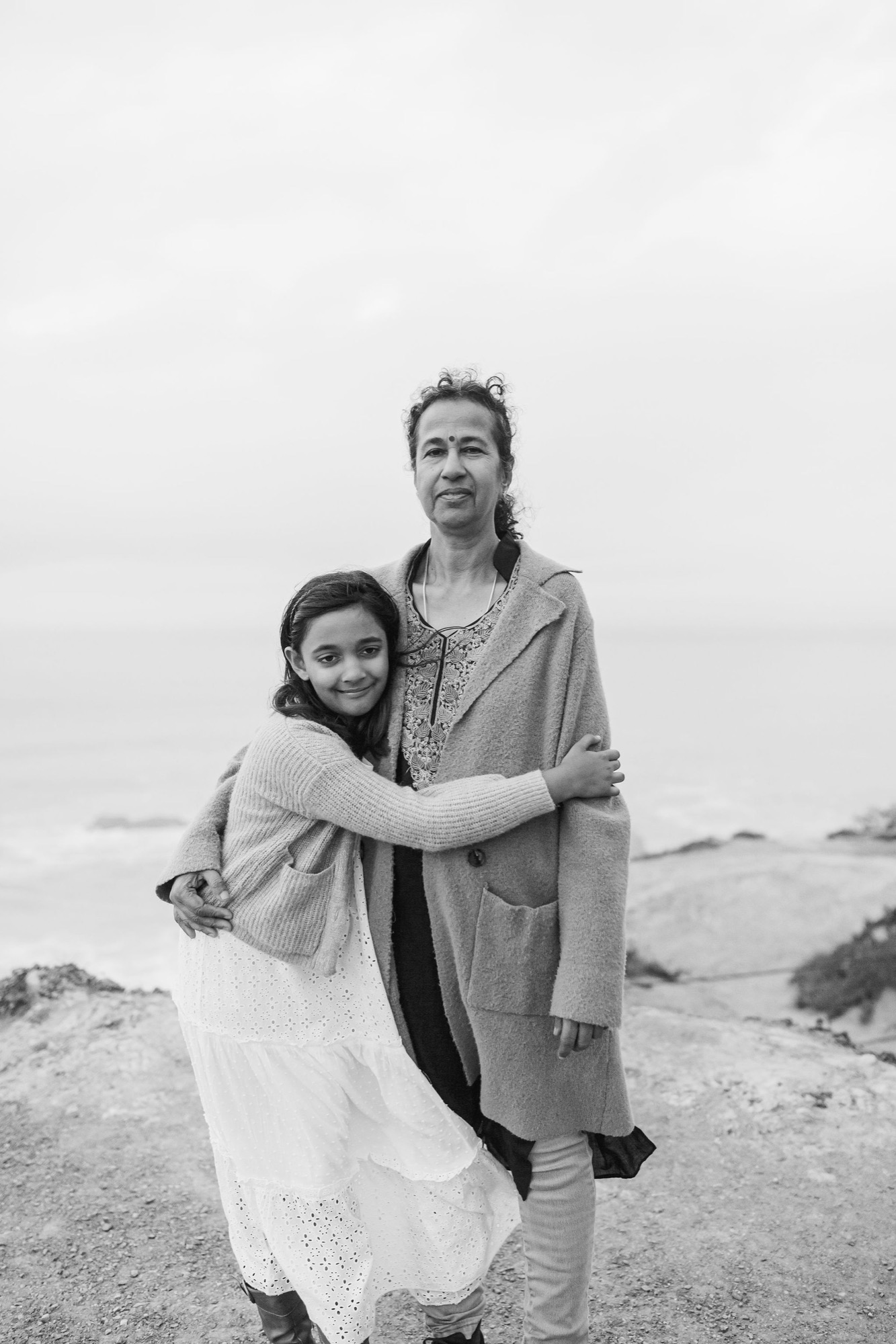 A woman and young girl embrace on a rocky coastal path, their warmth contrasting with the misty gray ocean and overcast sky behind them