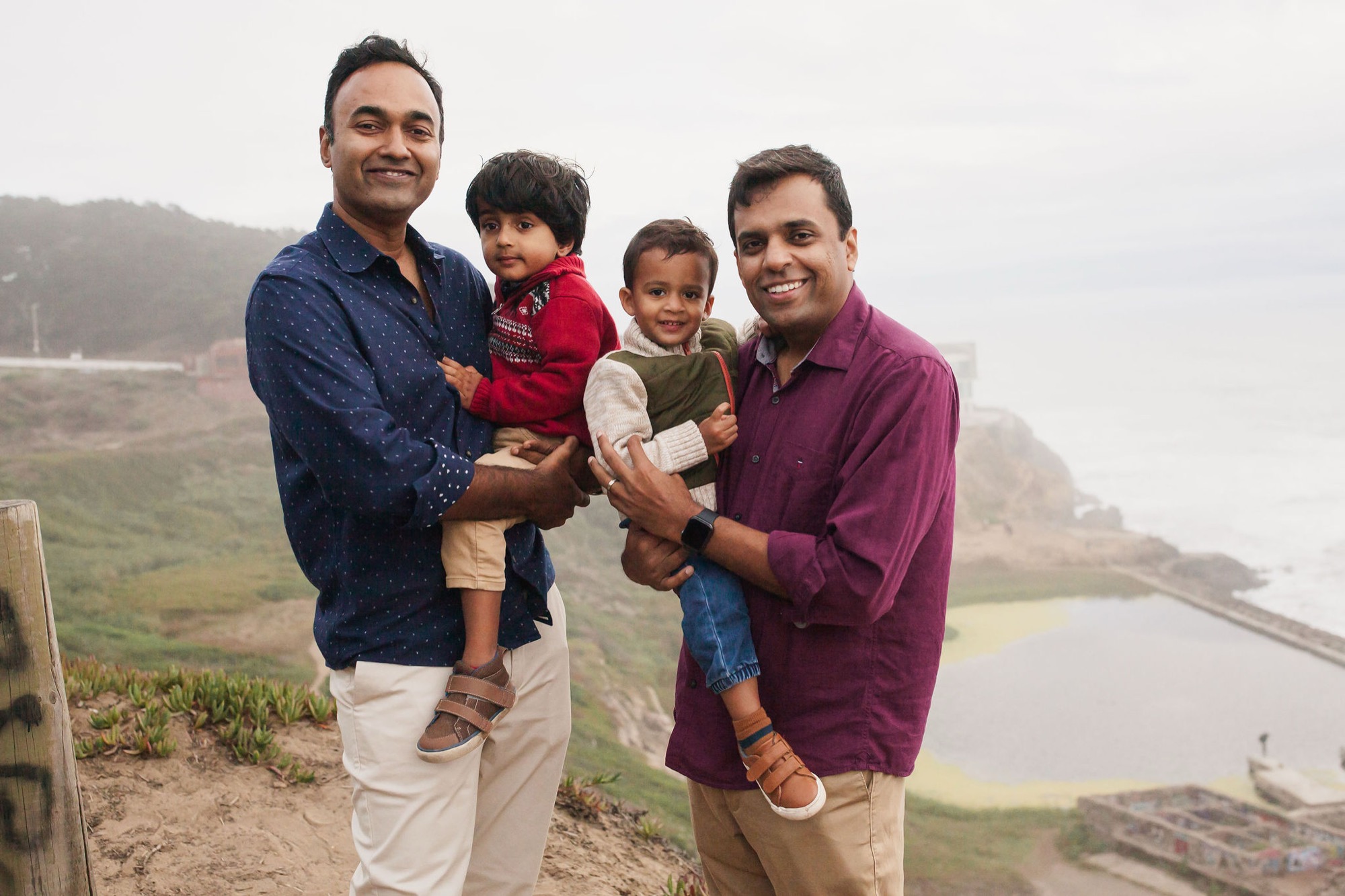 Two men hold young children on a misty hillside, smiling warmly at the camera with a coastal valley and water visible behind them