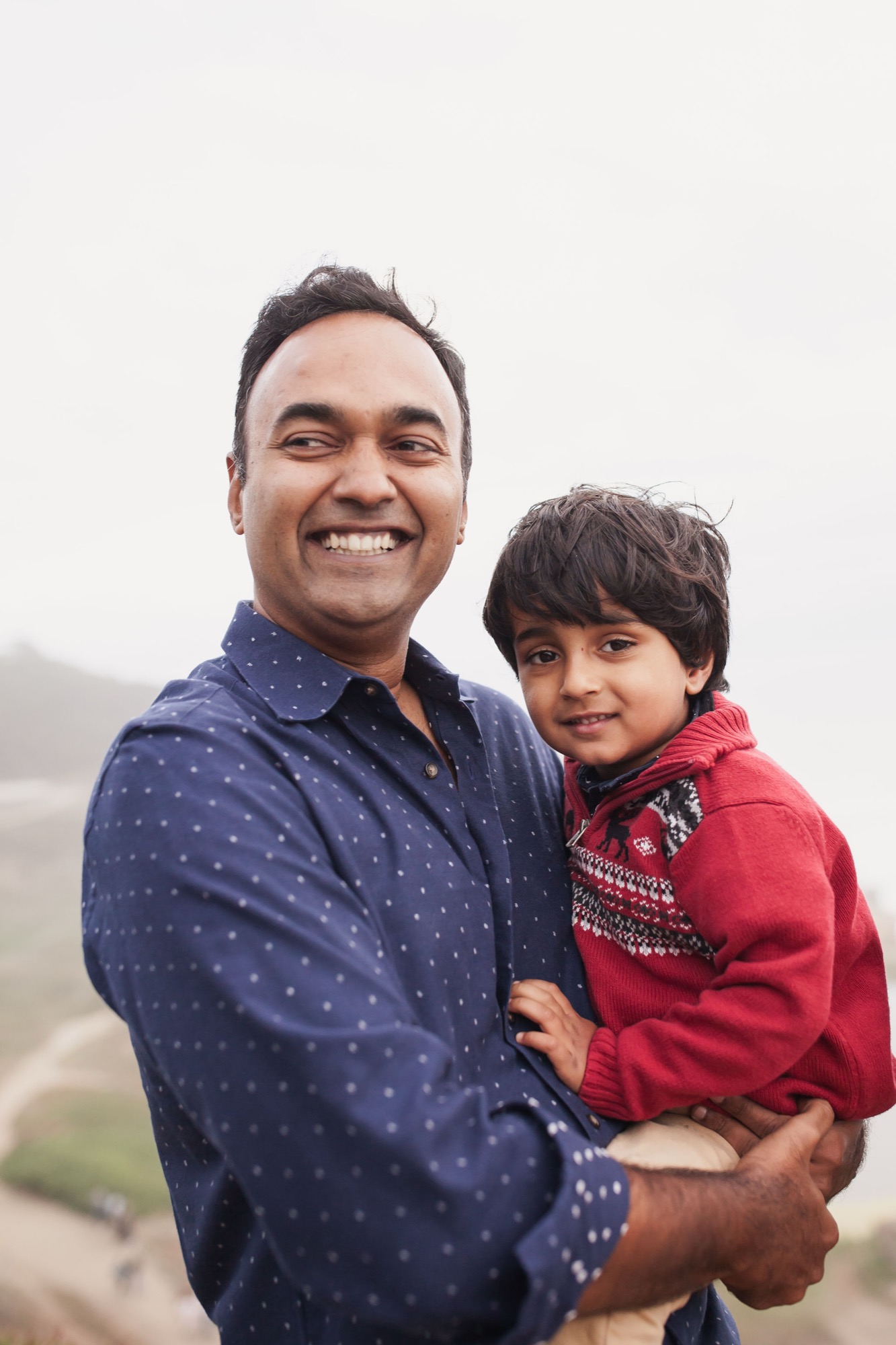 A beaming father holds his young son close against an overcast sky, both smiling warmly at the camera with blurred hillside backdrop