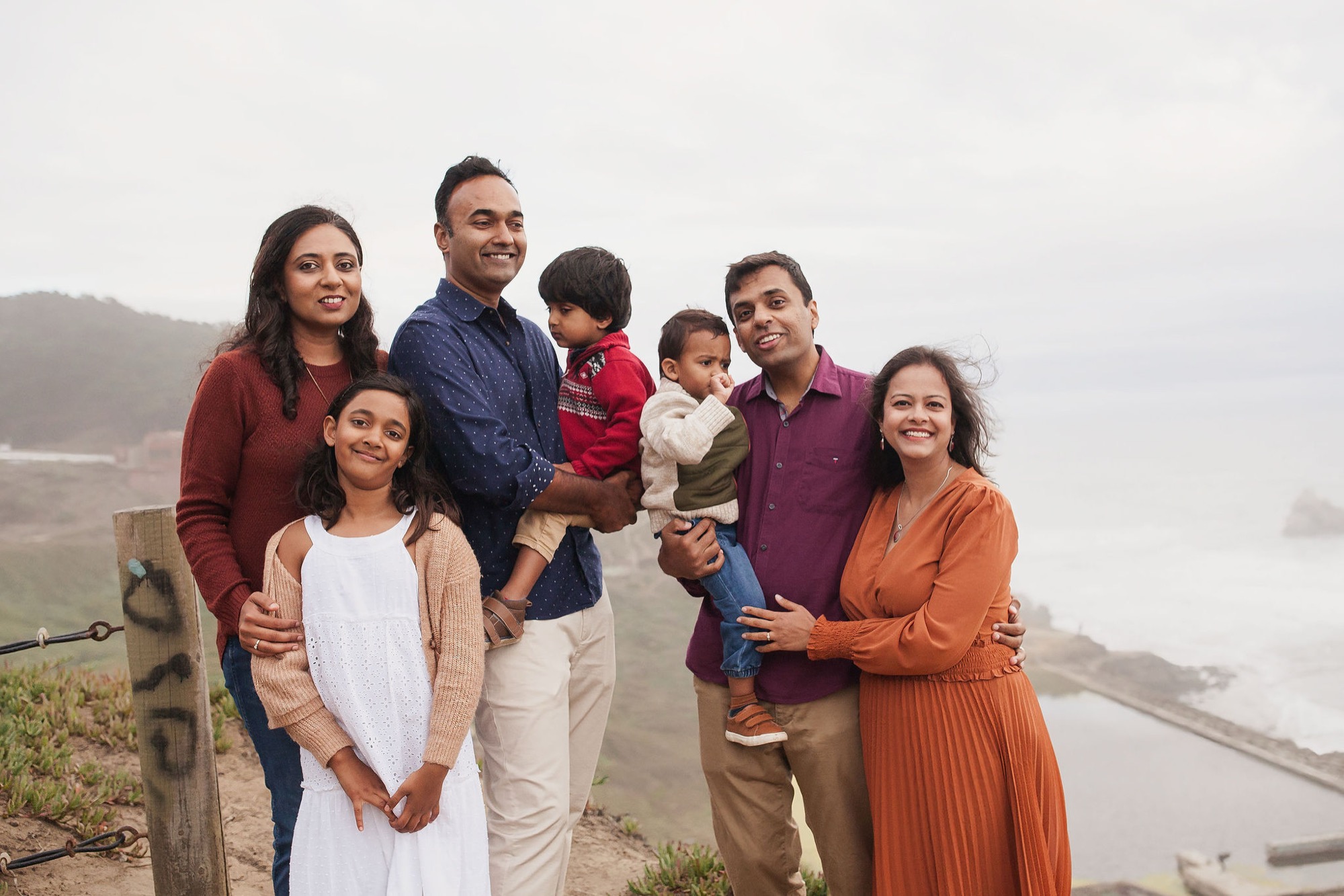 Six family members smile warmly at a misty overlook, their colorful autumn clothing contrasting against fog-shrouded hills and water below