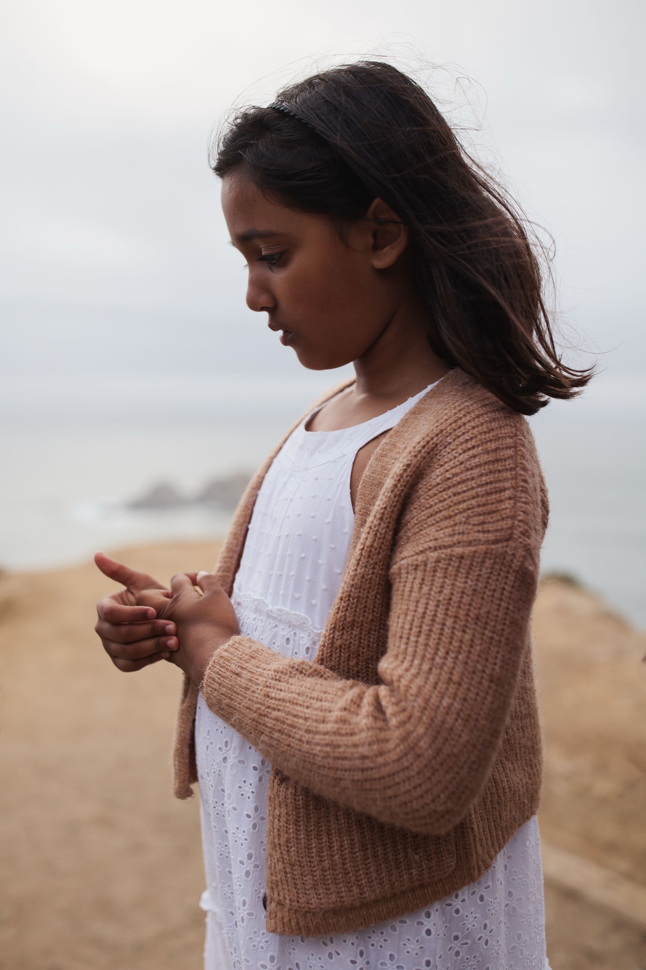 Woman in camel cardigan stands on windswept beach, gazing downward with contemplative expression as overcast sky meets distant shoreline