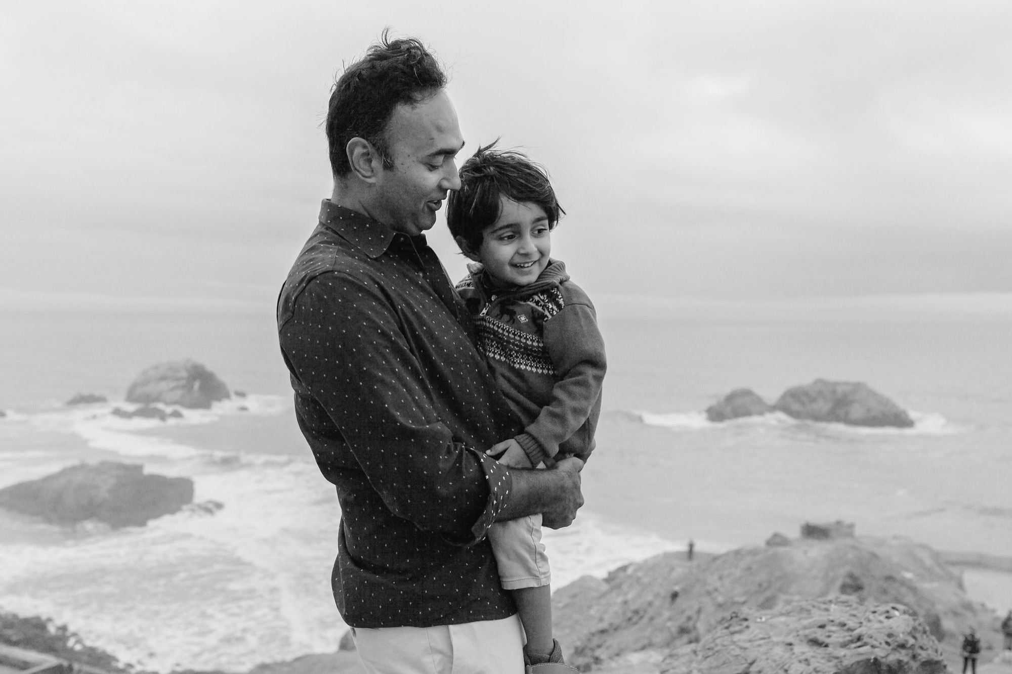 A father holds his young son at Land's End, both smiling as waves and rock formations blur softly behind them in overcast coastal light