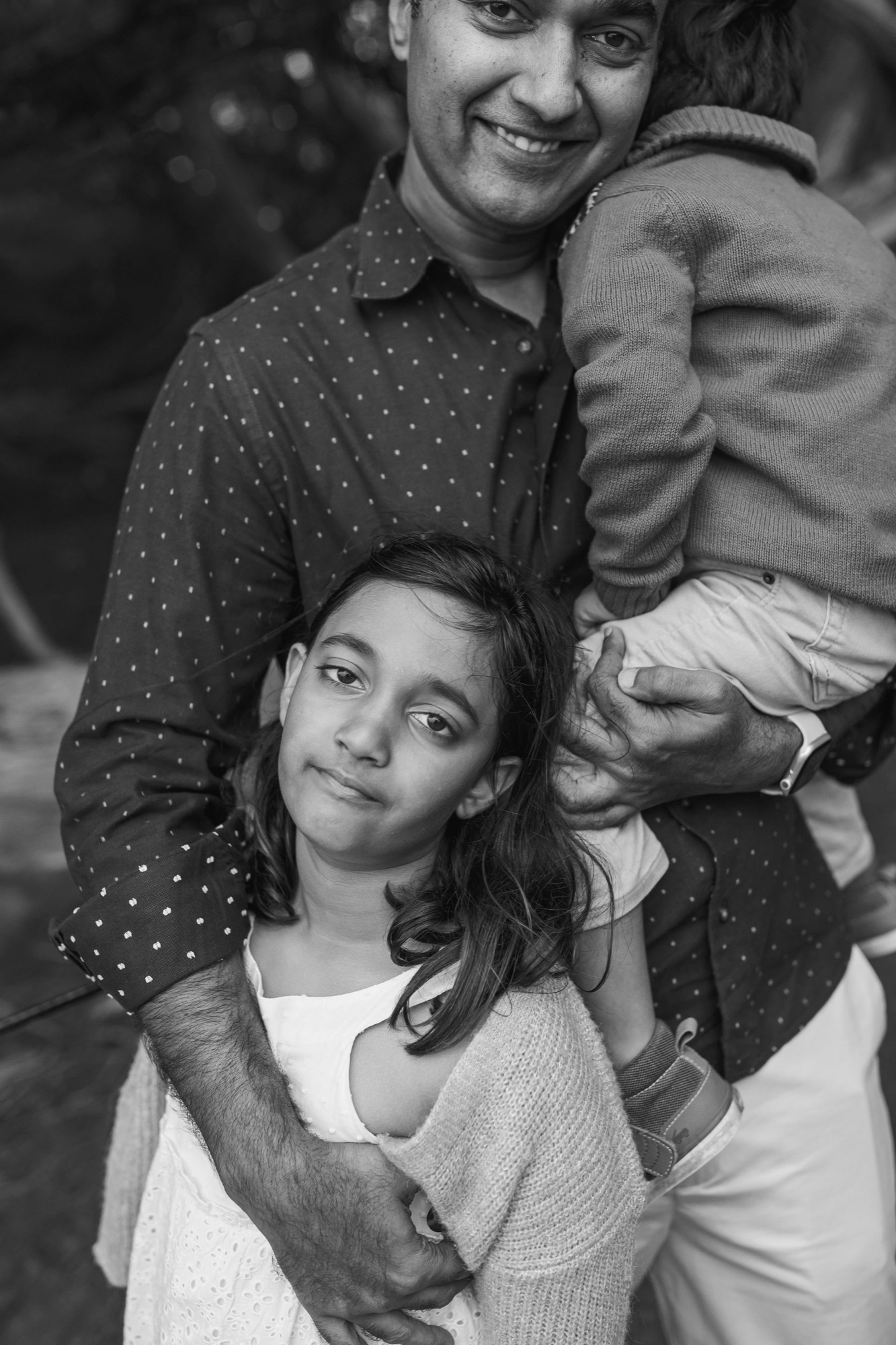 A father smiles broadly while holding his young son, with his older daughter standing close in front, all positioned together against a blurred natural background