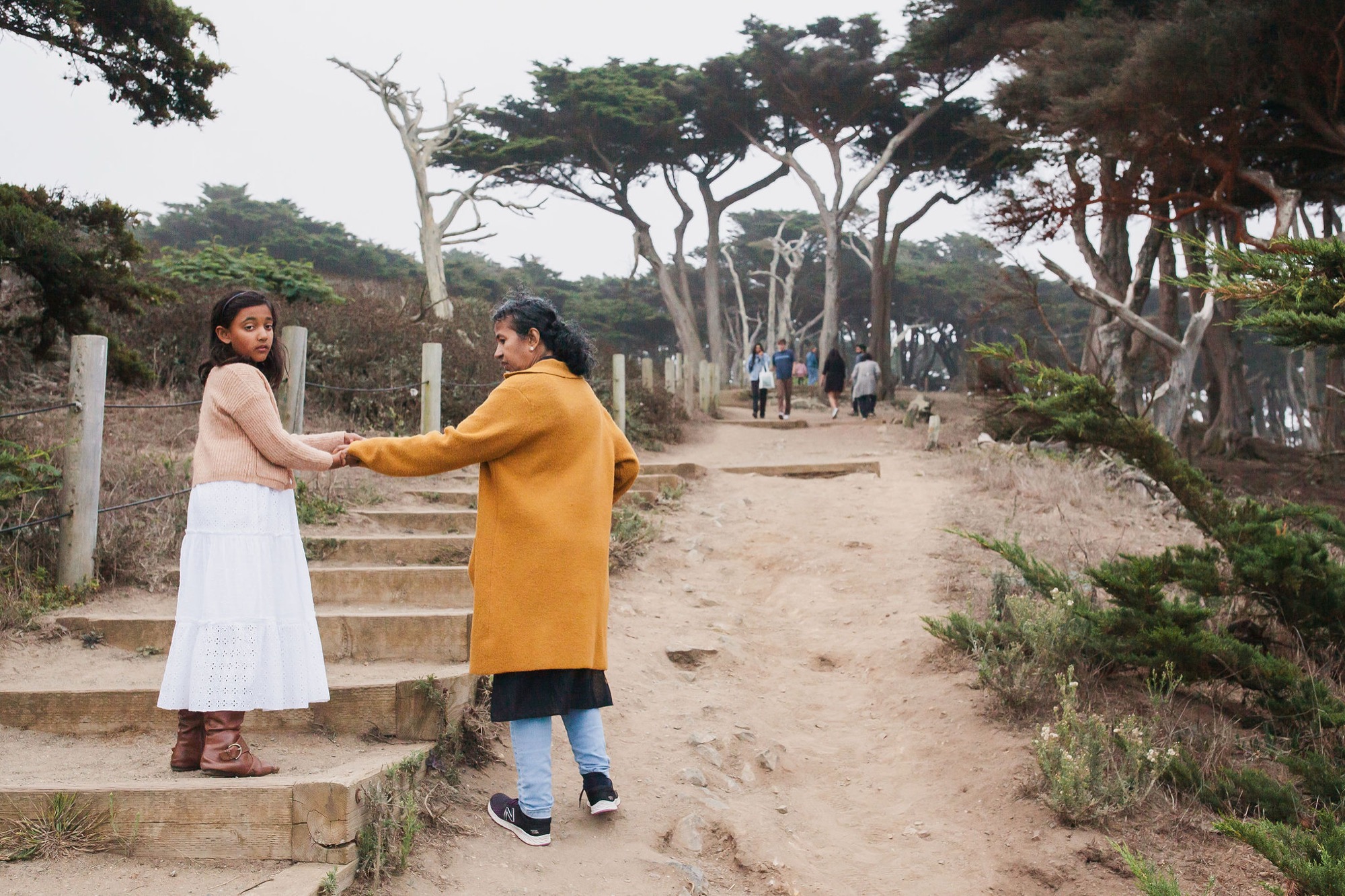 Two girls hold hands on a sandy trail beneath wind-sculpted cypress trees, turning back with genuine smiles toward the camera