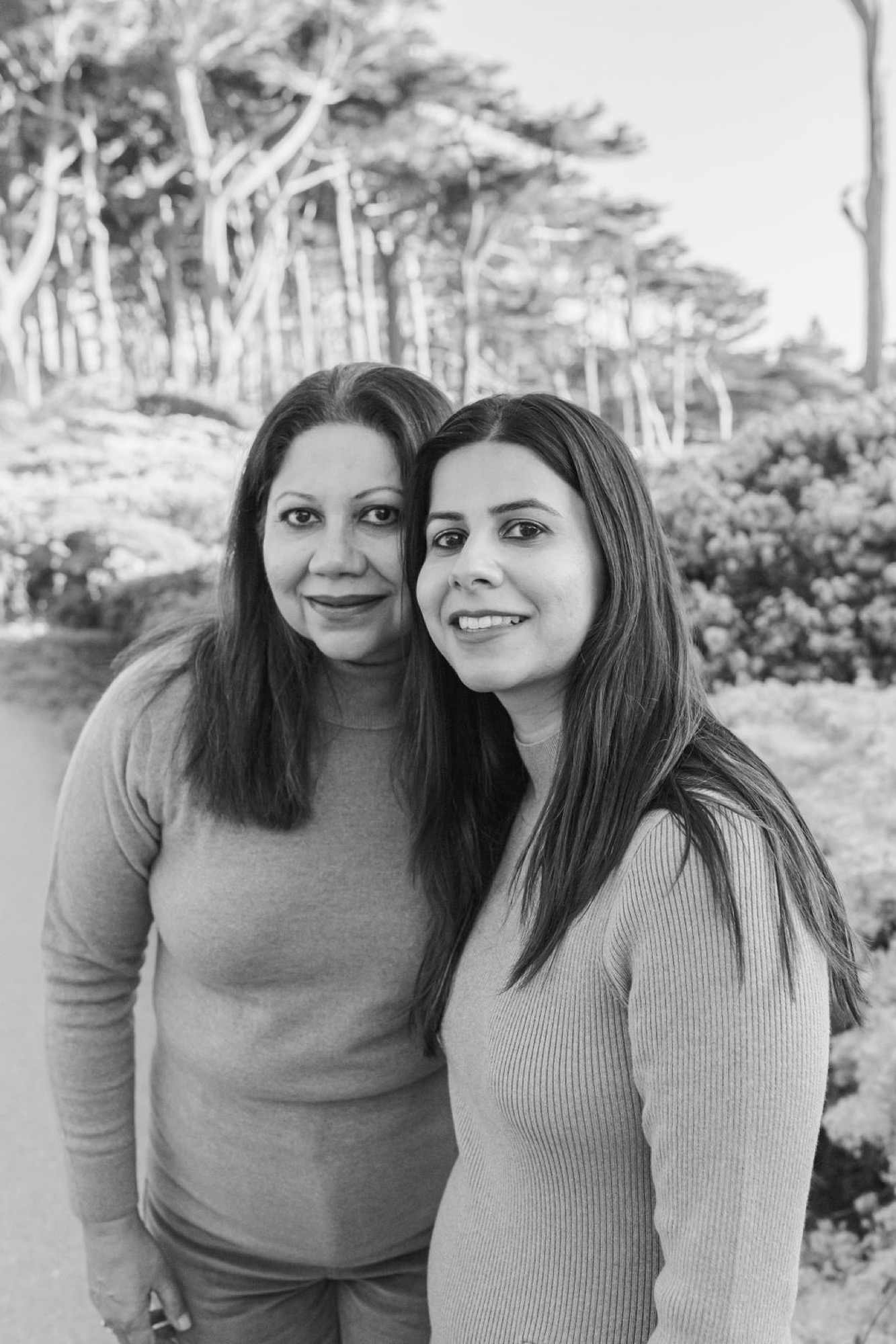 Two women with long dark hair stand close together against windswept coastal trees, both smiling naturally at the camera in soft natural light