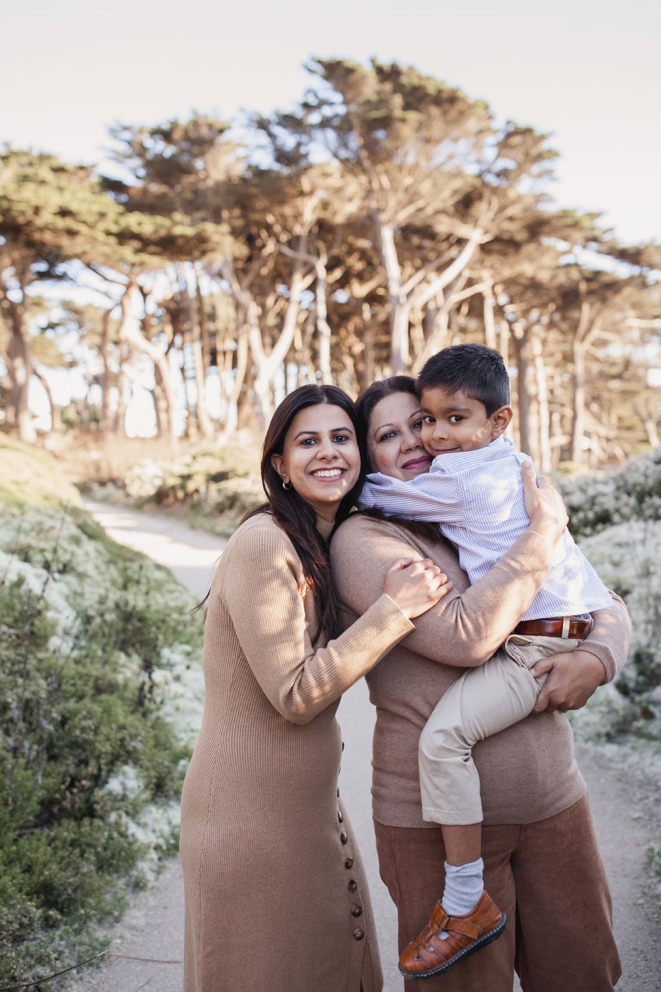 Two women and a young boy smile warmly on a coastal trail, with windswept Monterey cypress trees towering behind them in golden afternoon light