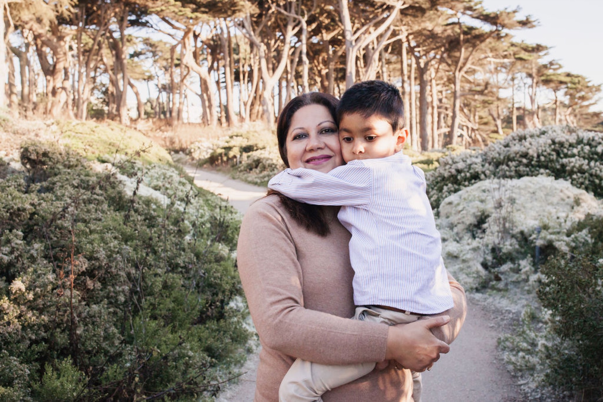 Mother holds her young son as he wraps his arm around her neck, both smiling warmly amid coastal cypress trees and shrubby vegetation