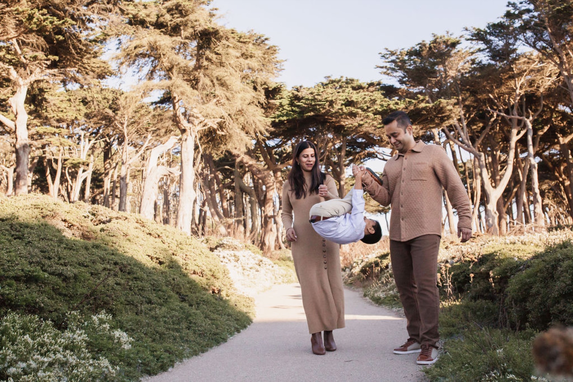 A father and mother walk together down a tree-lined path, playfully swinging their young child between them in the dappled sunlight