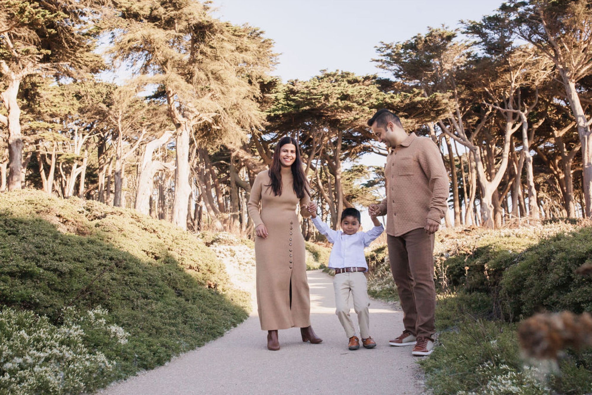 Young family walks hand-in-hand down a tree-lined path, their child swinging between them with genuine joy and connection