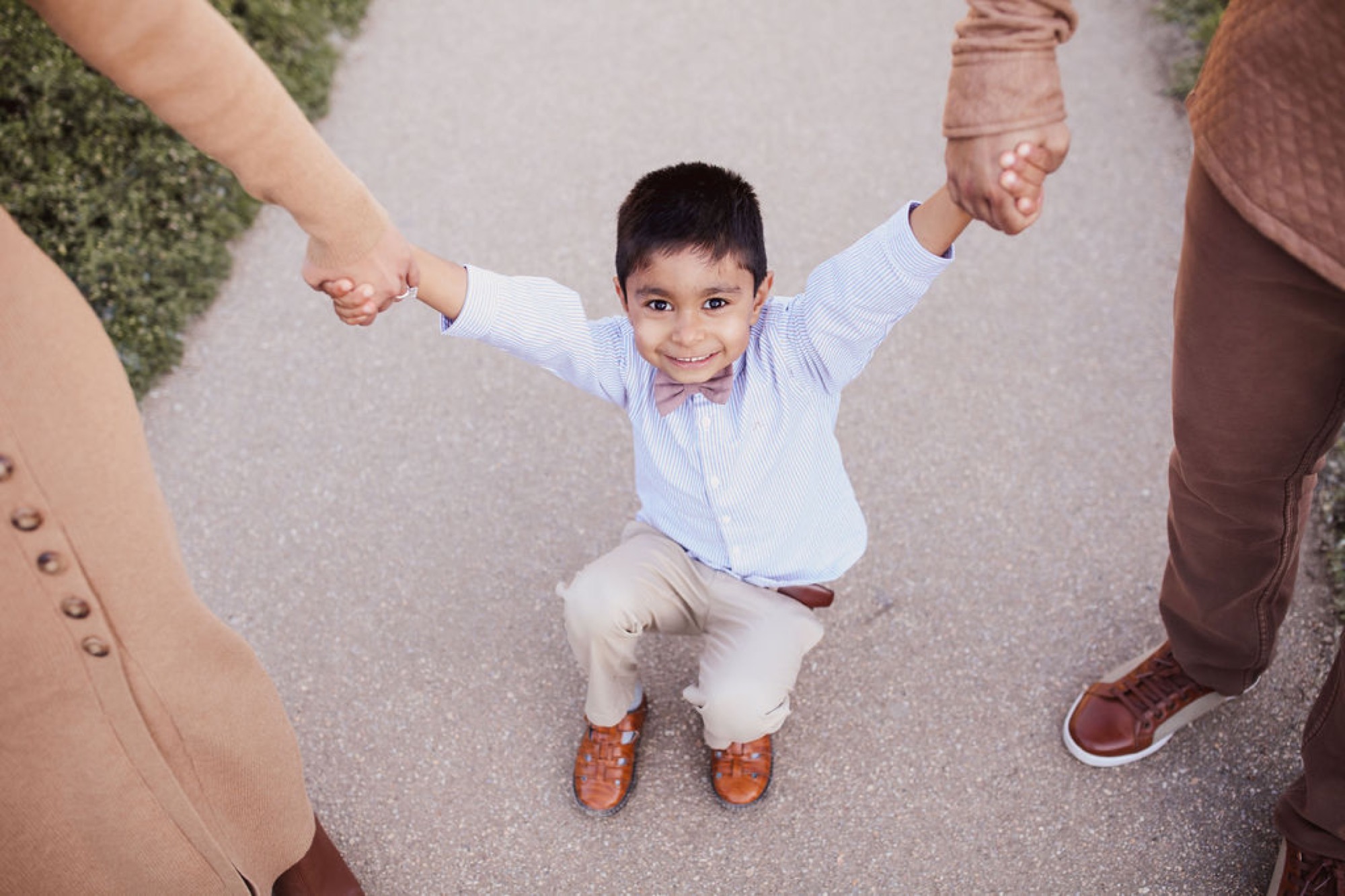 Young boy grins broadly while swinging between two adults on a concrete path, arms raised in joyful abandon