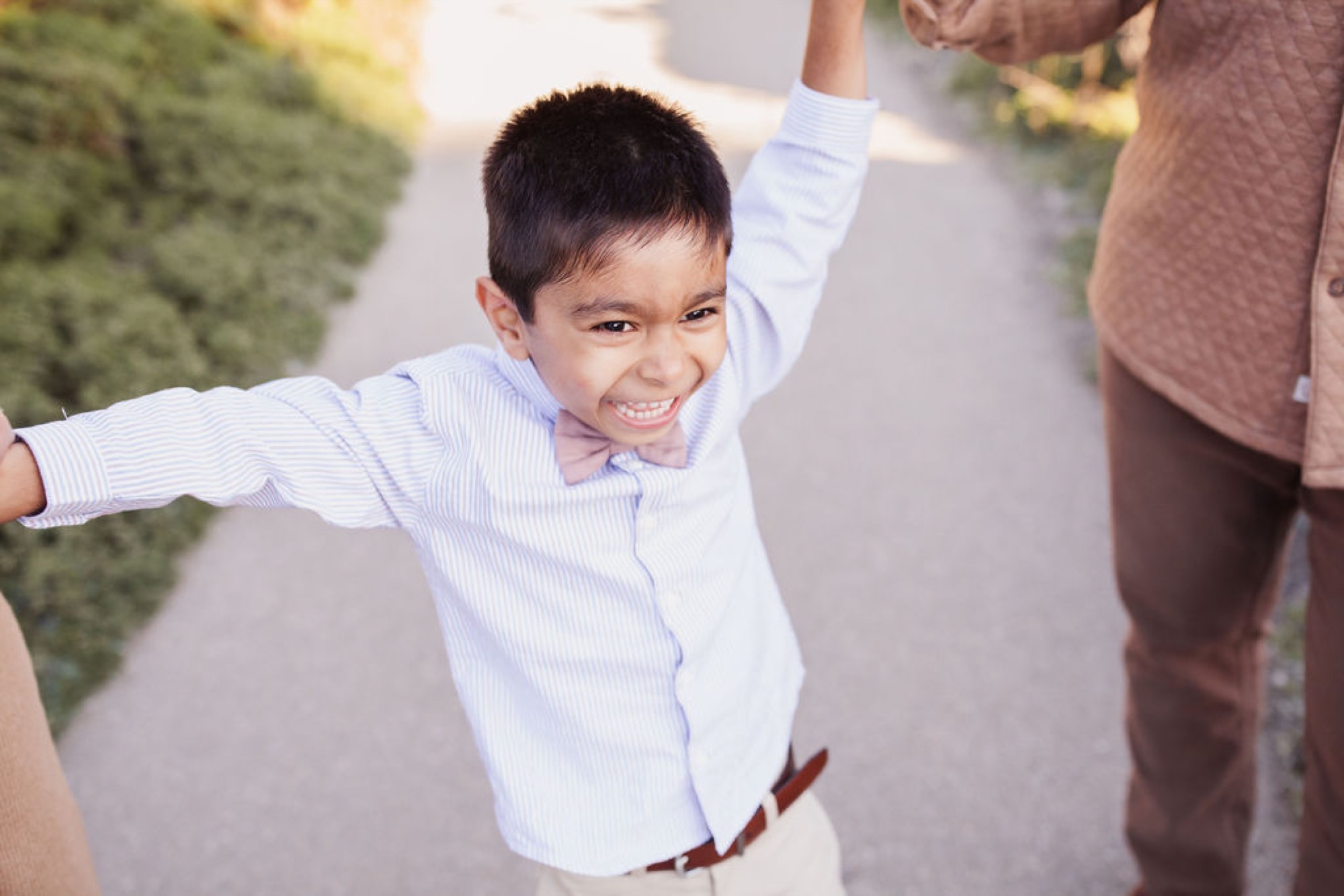 A young boy in a light blue shirt and pink bow tie swings joyfully between two adults along a sunny pathway, his radiant smile and outstretched arms capturing pure childhood delight