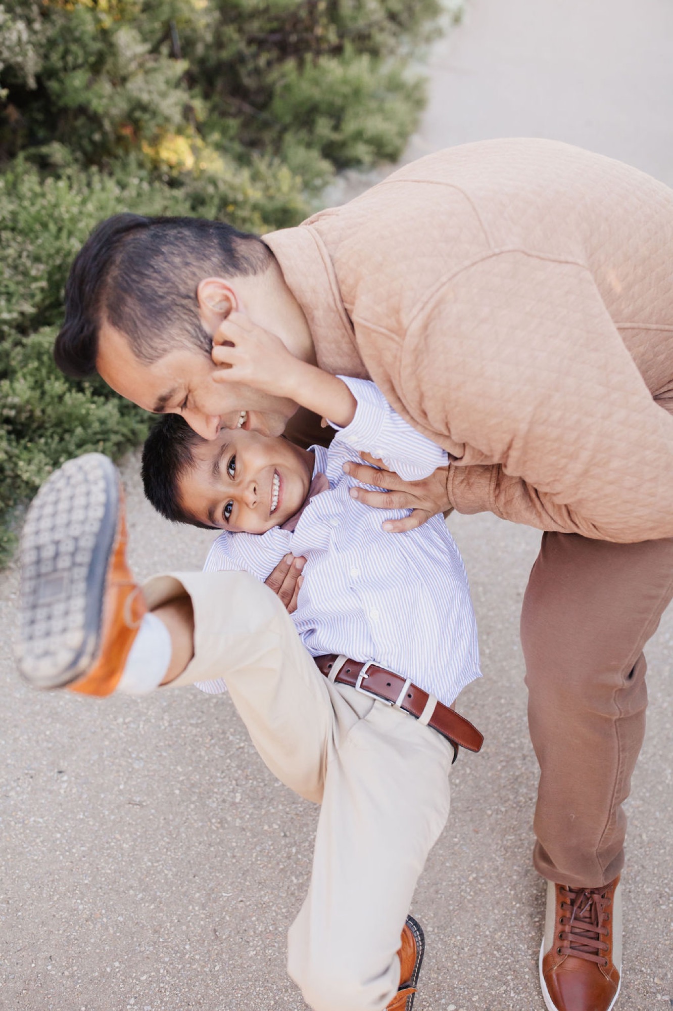 Father bends to kiss his young son's cheek while the boy laughs, one leg playfully kicked up on a sunny path