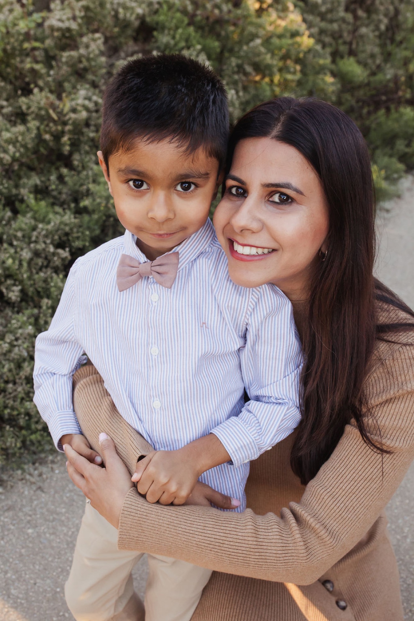 Woman holding young boy in striped shirt with pink bow tie, both smiling directly at camera against evergreen foliage backdrop