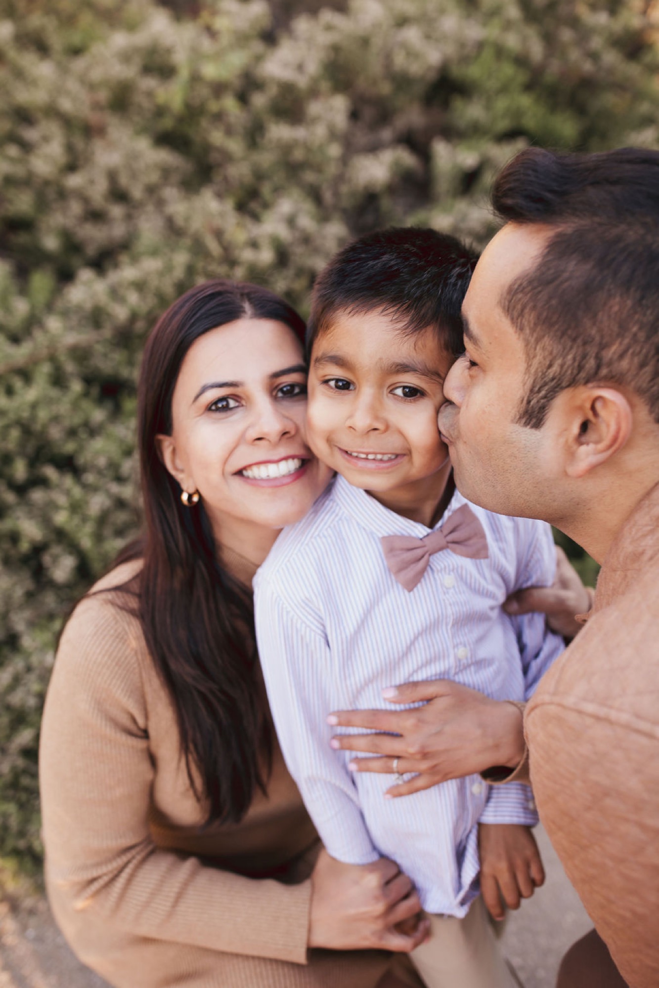 A smiling family of three poses together against a blurred green foliage backdrop, the child beaming between his parents in warm afternoon light