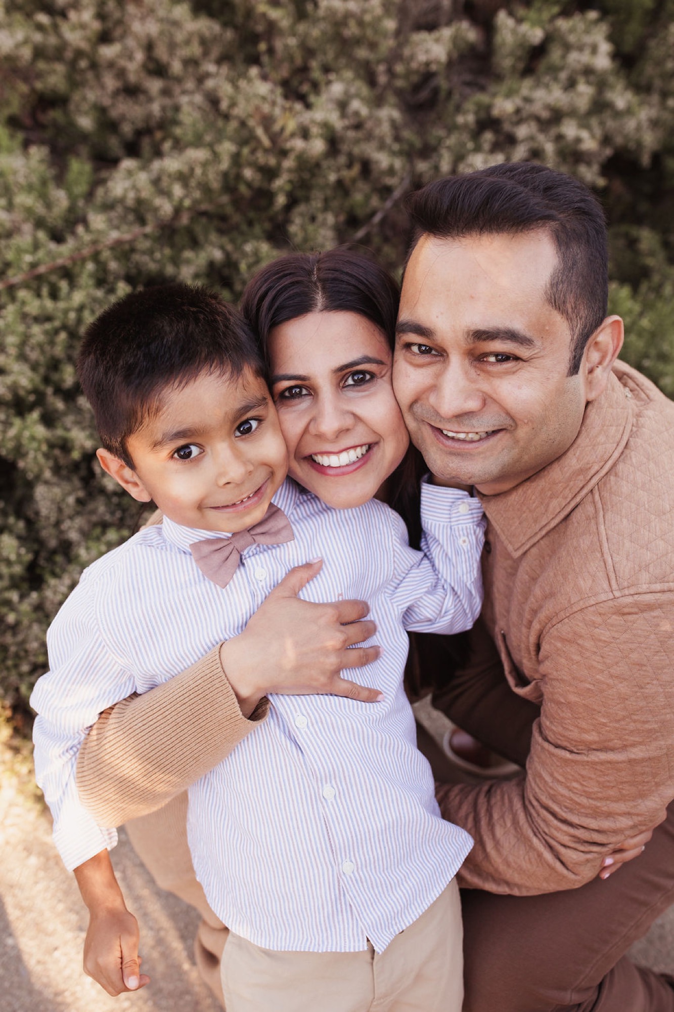 A father holds his young son while the mother embraces them both, all three smiling warmly against a backdrop of weathered coastal vegetation