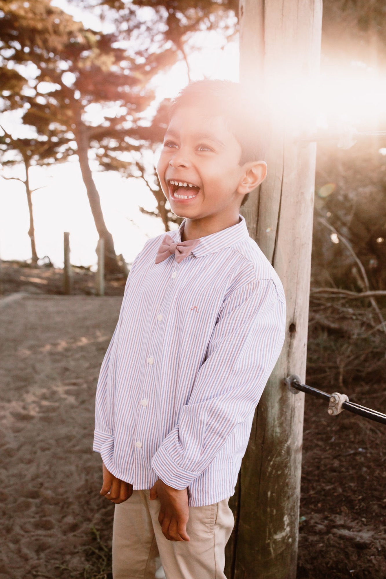 A young boy in a lavender striped shirt and pink bow tie laughs joyfully against a weathered wooden post, backlit by golden sunset light