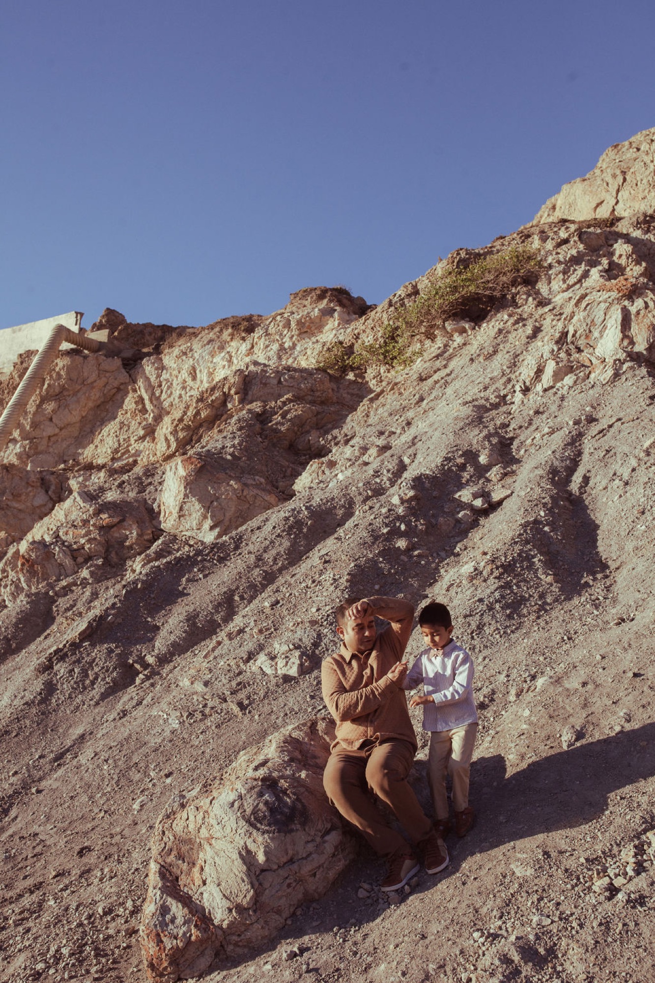 A woman in a tan jacket crouches beside a young child on a sun-bleached rocky slope, her hand gently guiding his arm
