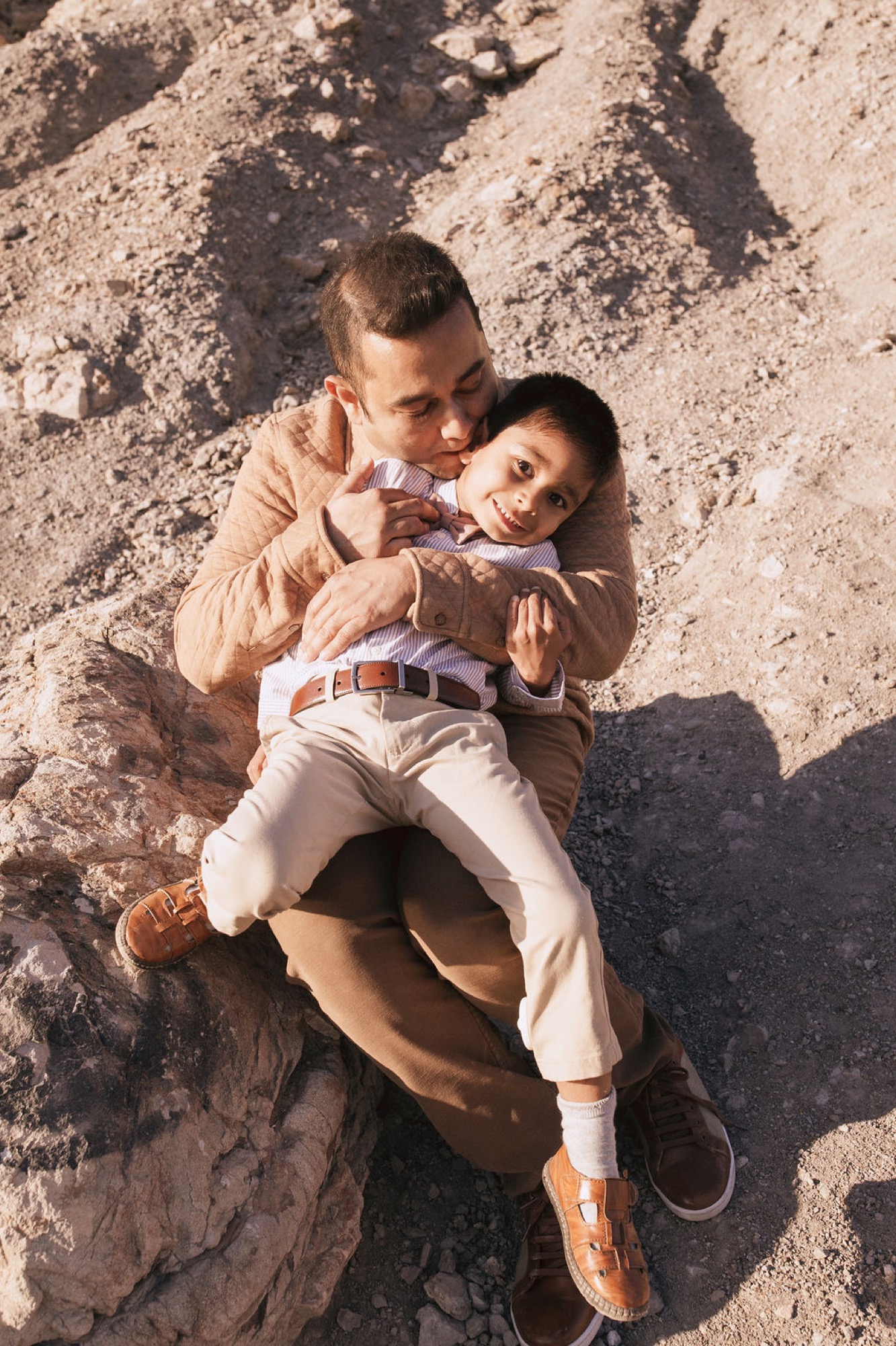 Two young boys lie on sun-dappled dirt, one embracing the other from behind with genuine affection and bright smiles
