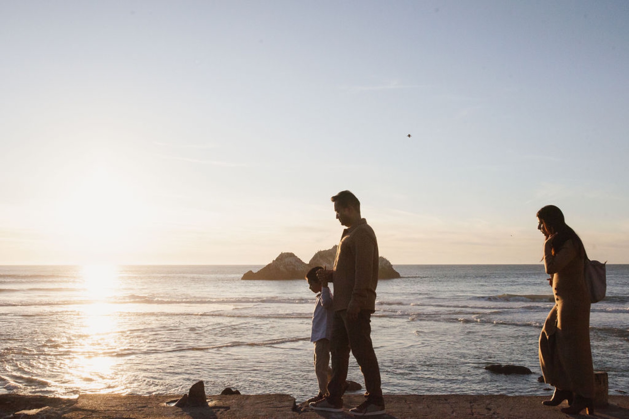A father holds his young child while standing on the beach at sunset, with his partner nearby watching the golden light reflect off the water