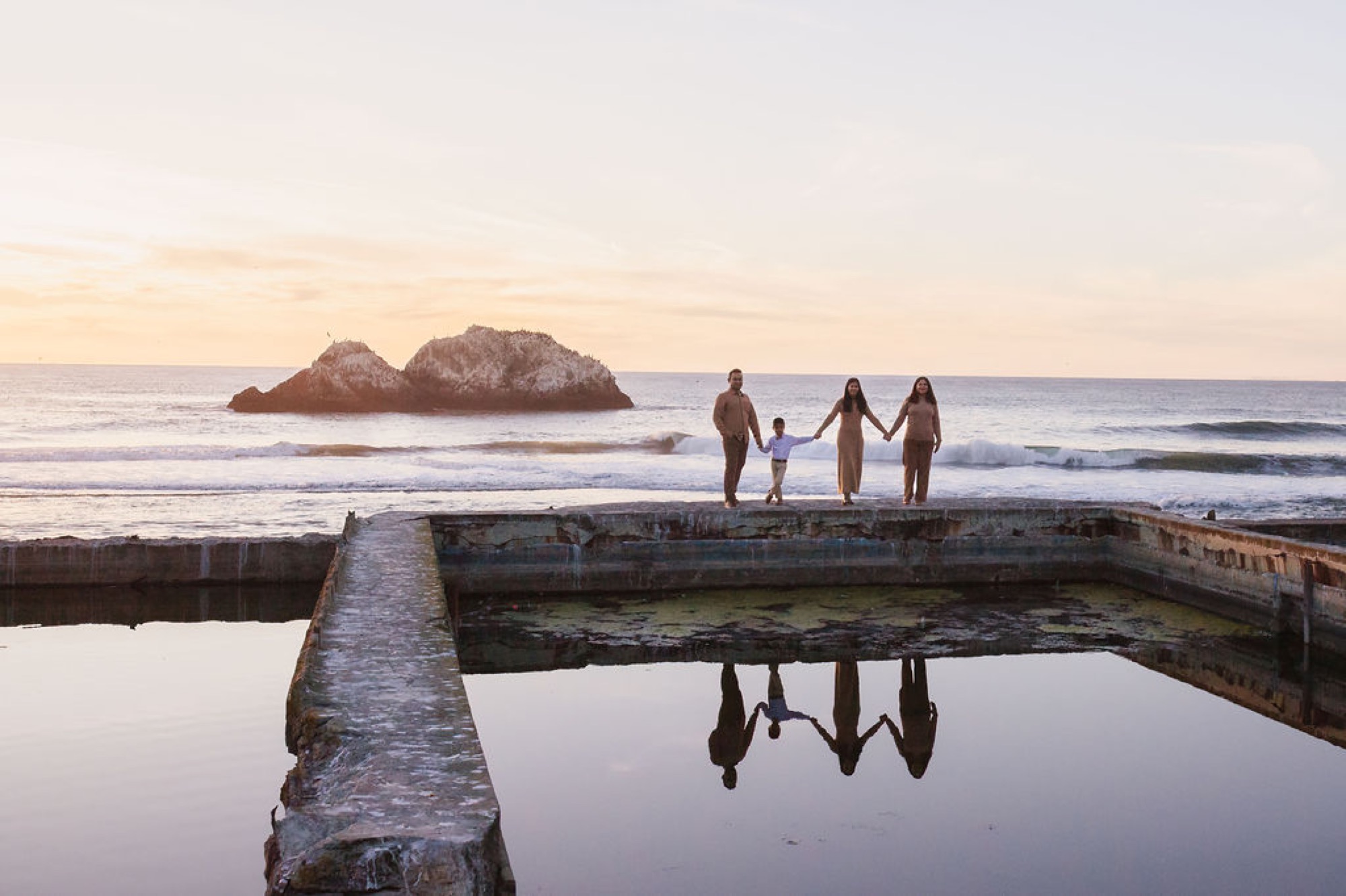 Four figures in swimwear stand holding hands on a weathered coastal pool structure, their reflections mirrored below as golden evening light bathes the rocky headland beyond